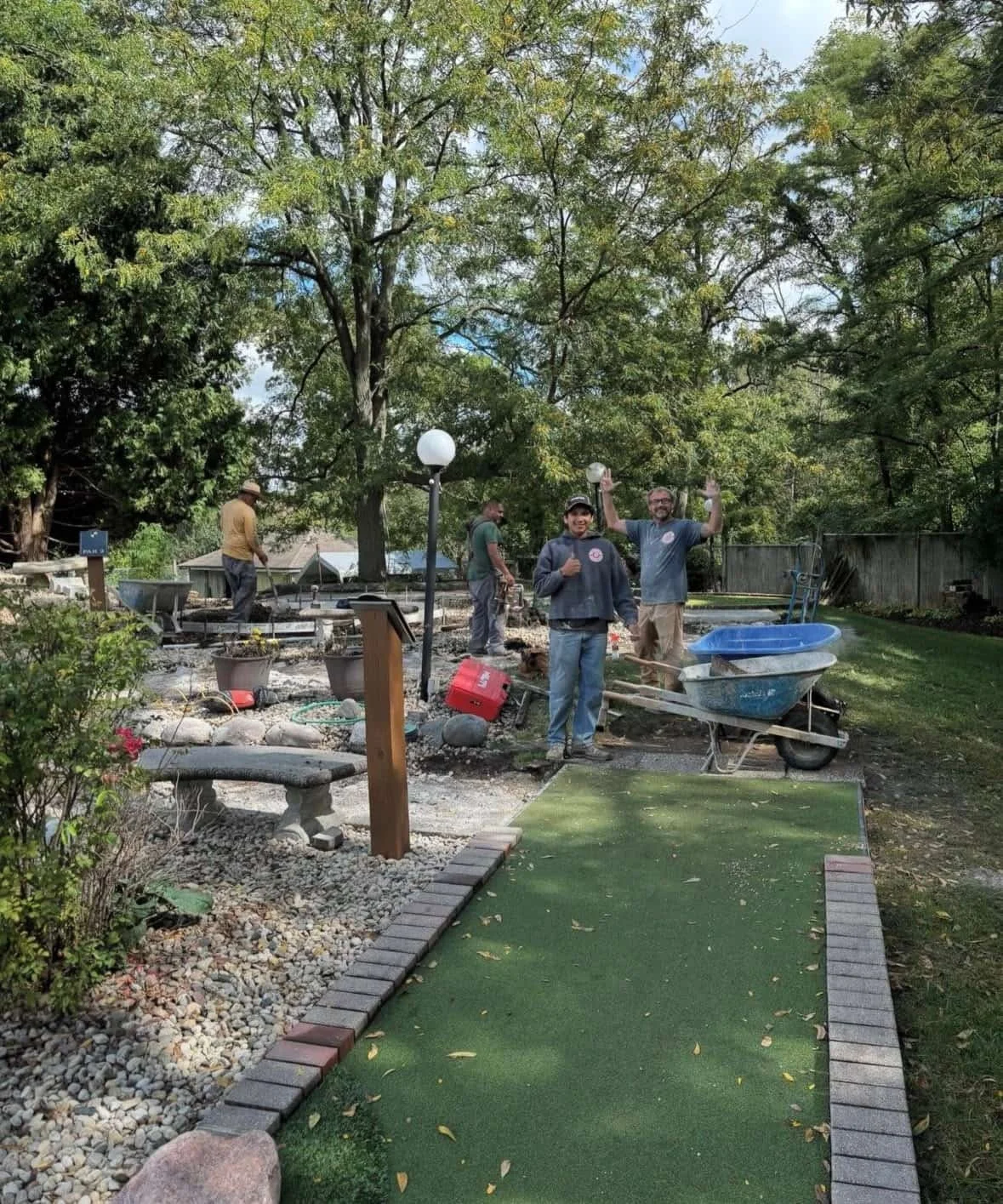 Crew working on landscaping or construction project in a backyard with trees, a mini golf course, and a wheelbarrow.
