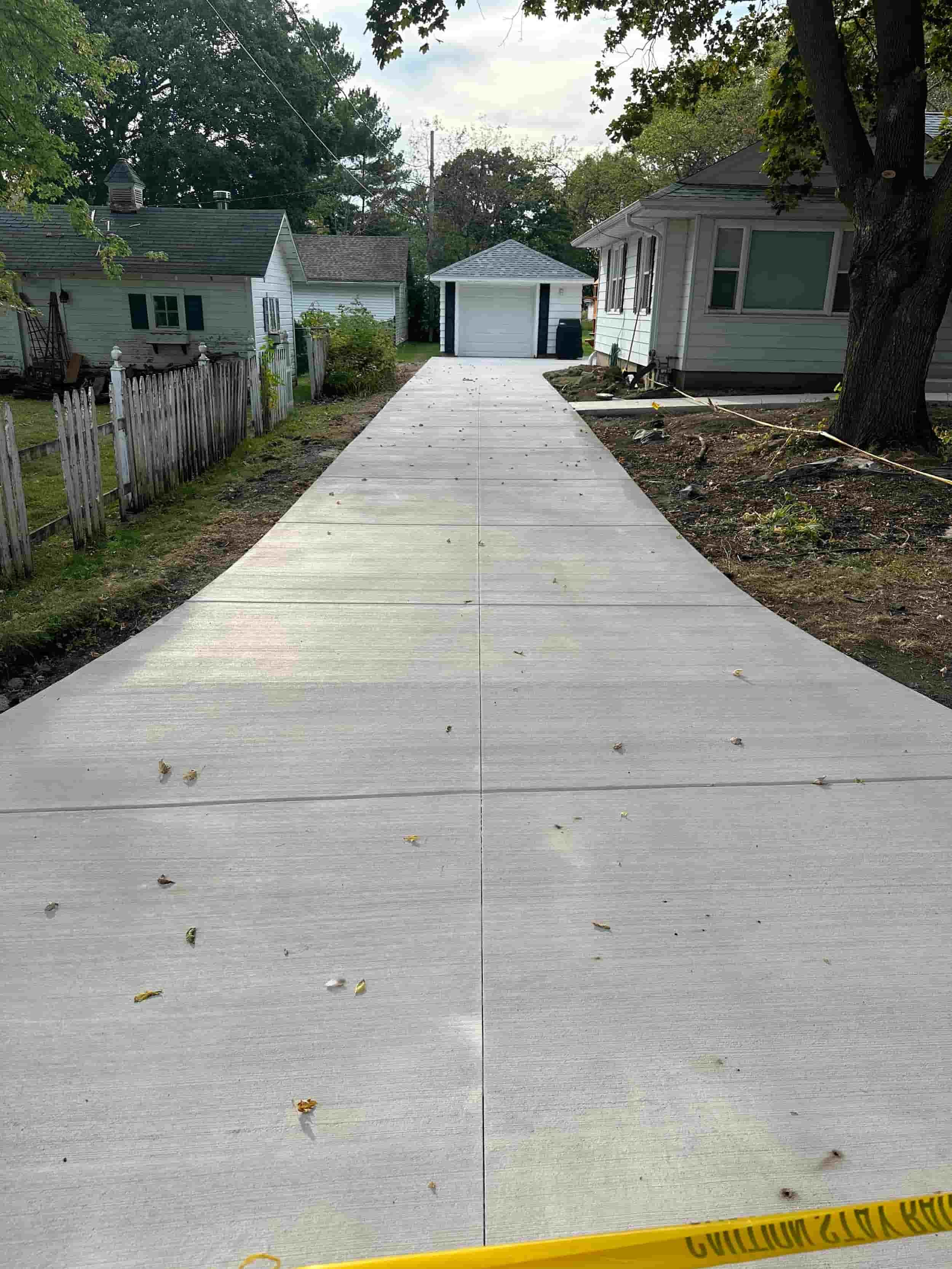 Newly paved concrete driveway leading to a detached garage, with houses and trees on either side, some scattered leaves on the driveway, and a yellow warning tape at the bottom.