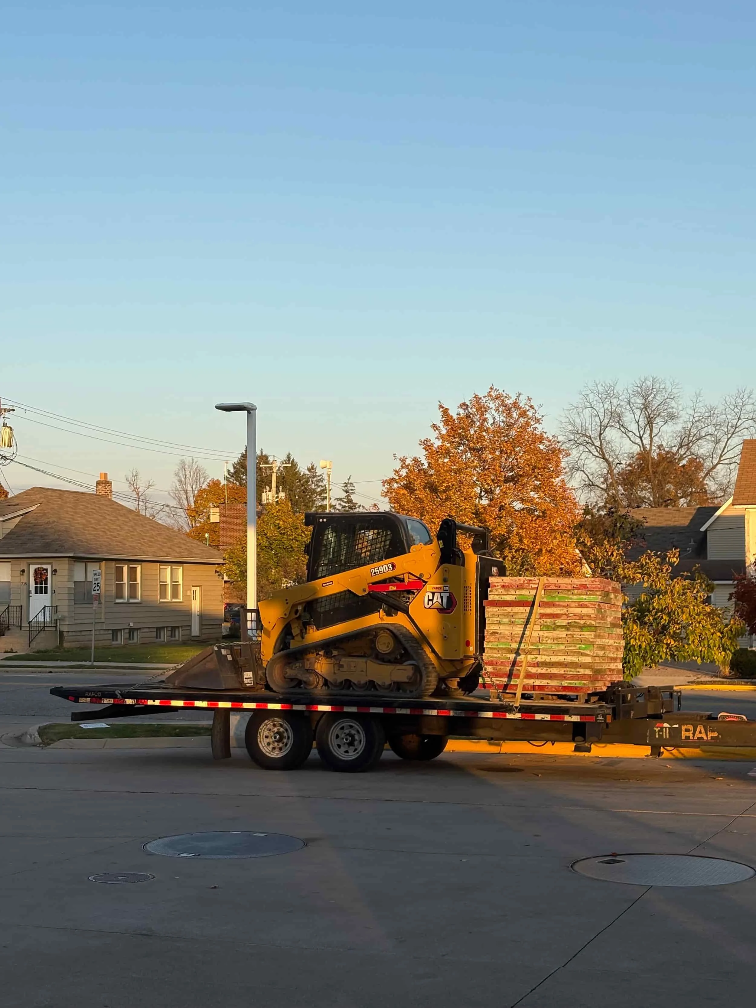 A yellow CAT compact track loader on a flatbed trailer, parked on a street with houses and autumn trees in the background.