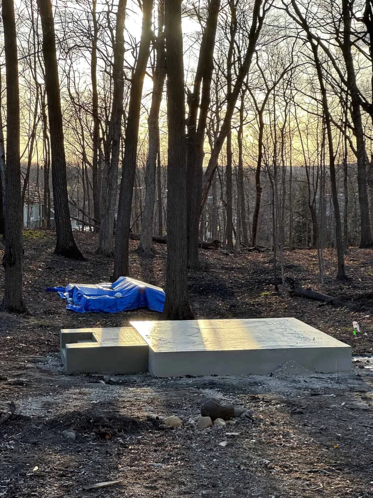 A newly built concrete walkway or platform in a wooded area with tall trees and a pile covered with a blue tarp in the background during sunset or sunrise.