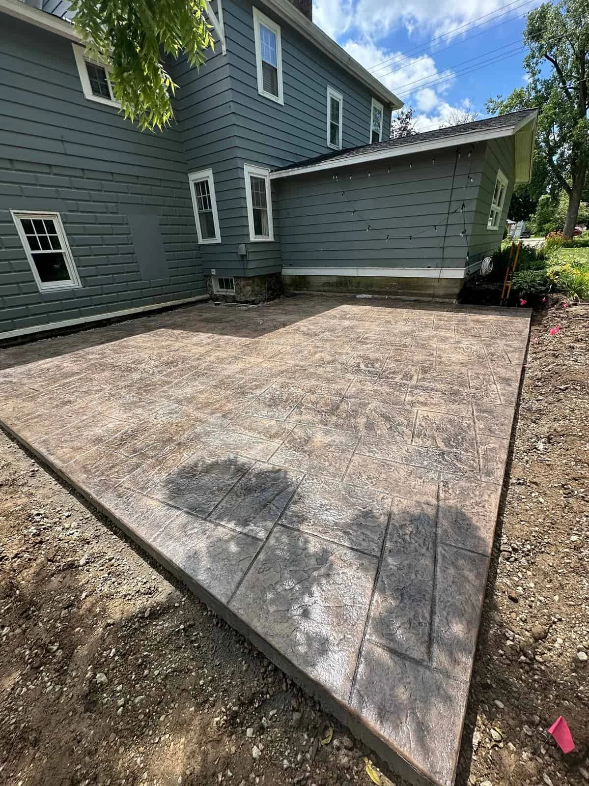 Newly installed stamped concrete patio in the backyard of a blue house, with some shadows and a small garden area to the side.