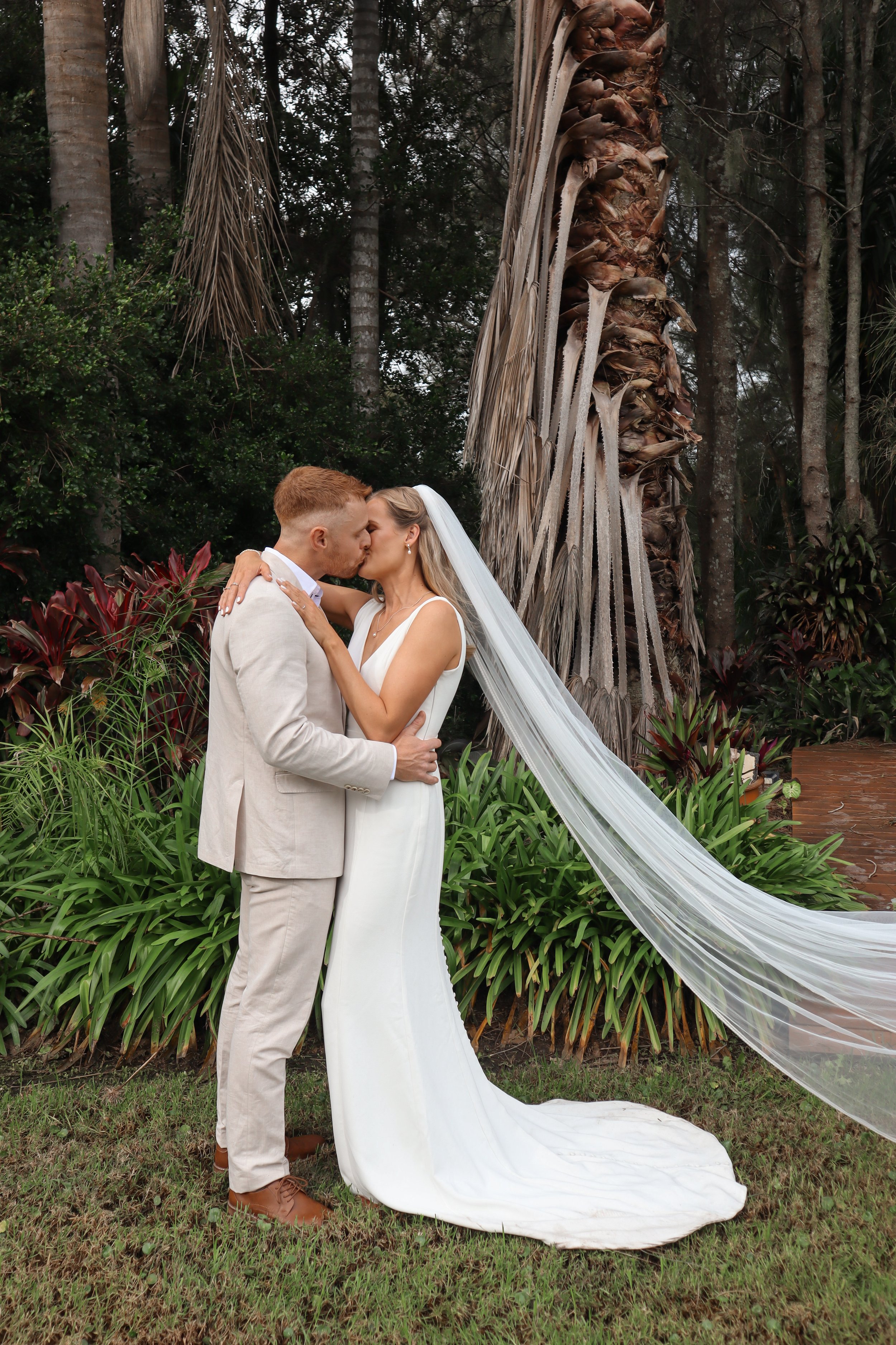 A bride and groom sharing a kiss outdoors, with the bride's long veil blowing in the wind. The bride is wearing a white wedding dress, and the groom is dressed in a light-colored suit. They are standing on grass in front of lush green foliage and tall palm trees.