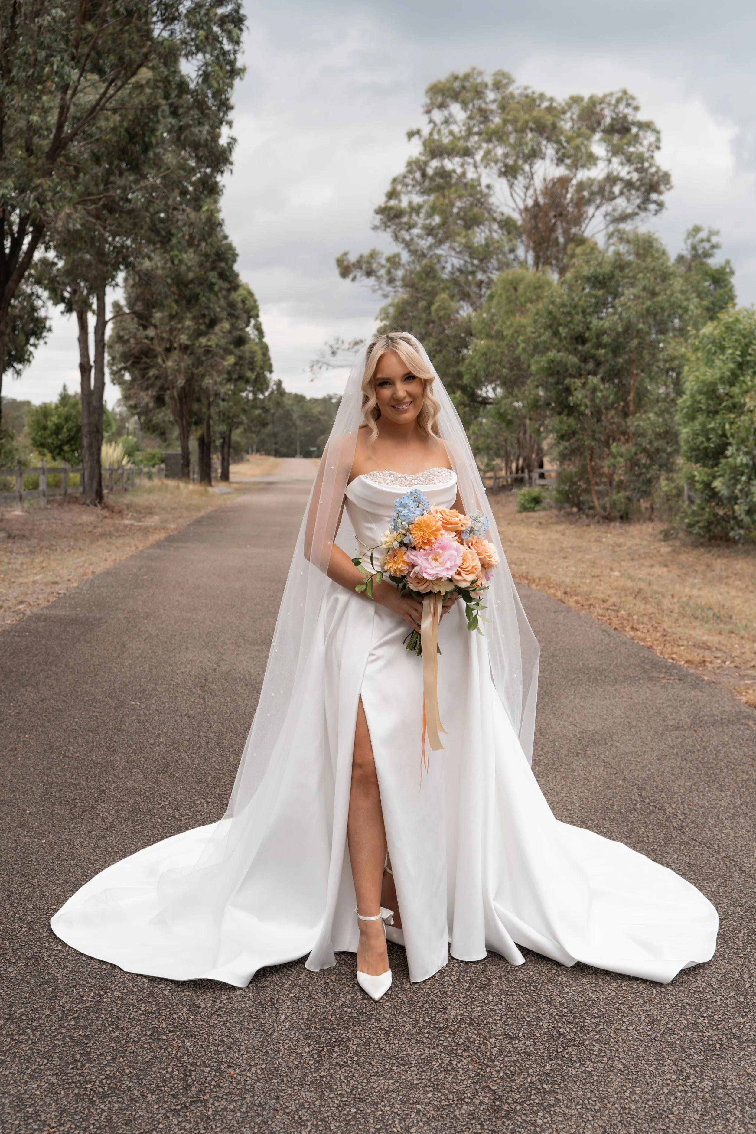 A bride in a white wedding dress holding a colorful bouquet, standing on a paved road surrounded by trees and greenery.