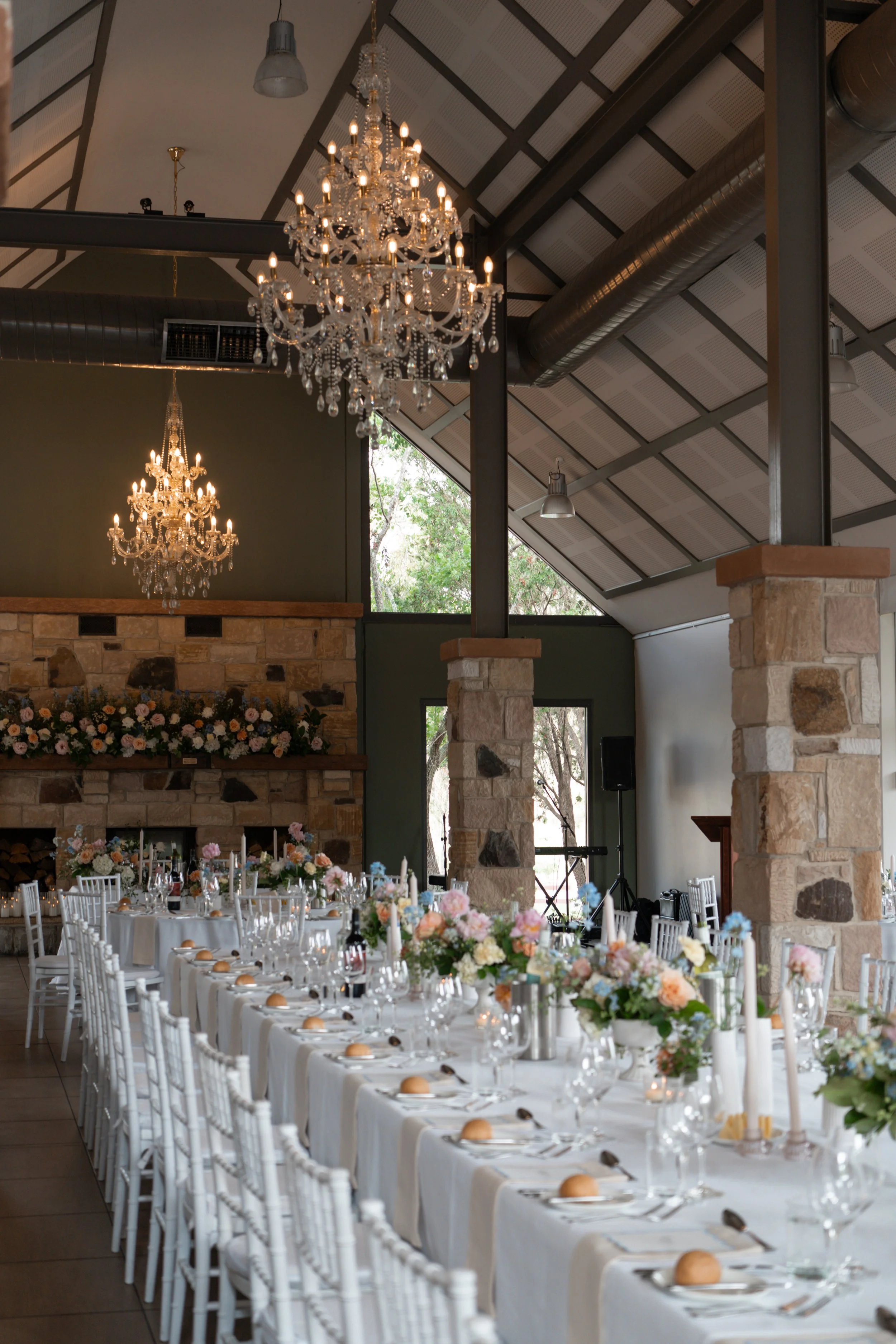 Elegant indoor wedding reception with long table set with white tablecloths, floral centerpieces, candles, and plates with bread rolls. Crystal chandeliers hang from the vaulted ceiling with exposed beams and ductwork. Large windows reveal trees outside.