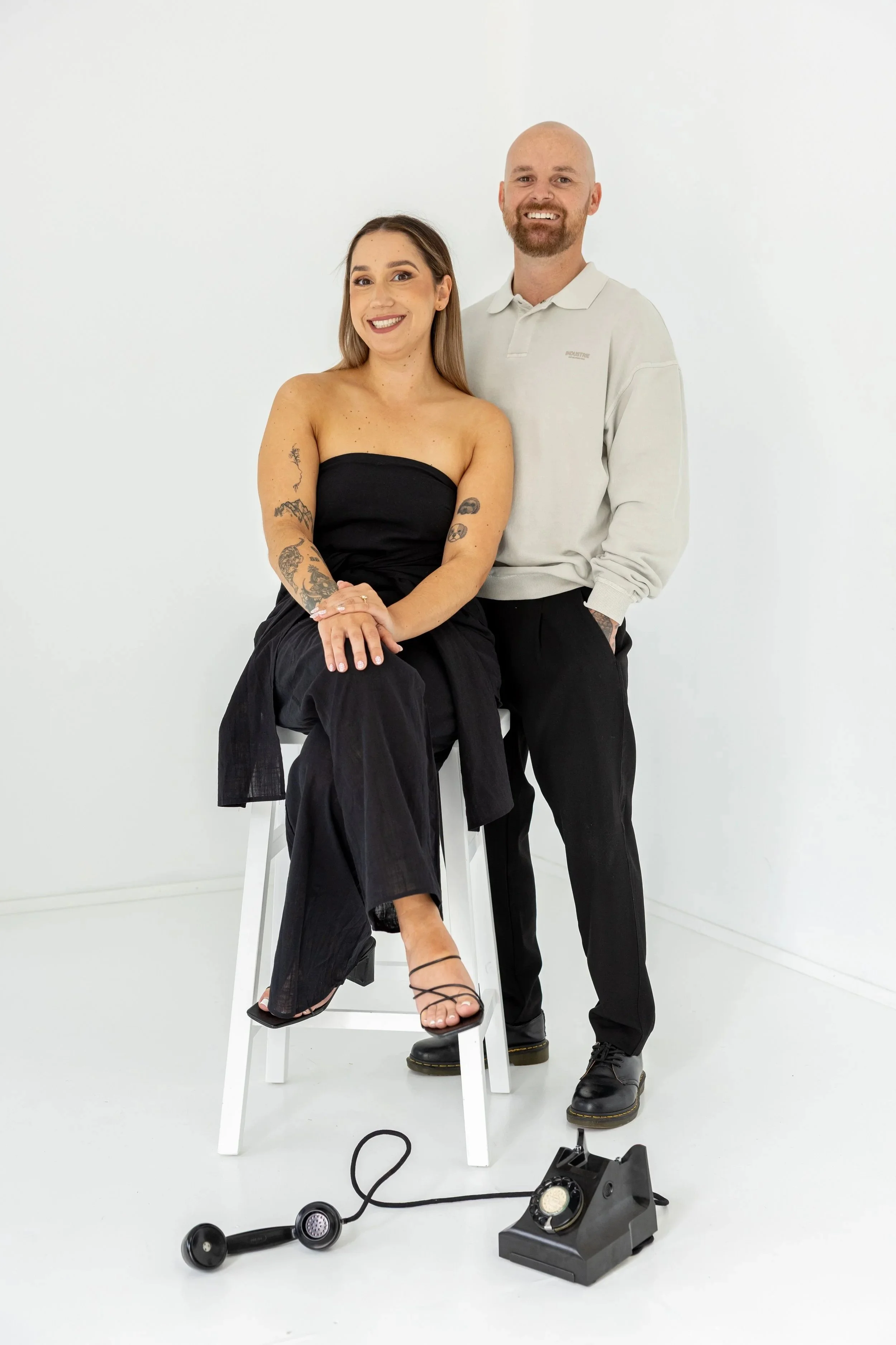 A smiling woman in a strapless black top and black pants, with tattoos on her arms, is sitting on a white stool. Next to her is a man with a beard and a shaved head, wearing a light-colored hoodie and black pants, standing with his hand in his pocket. Items including a vintage black rotary telephone and a black telephone receiver are on the floor in front of them, against a plain white background.