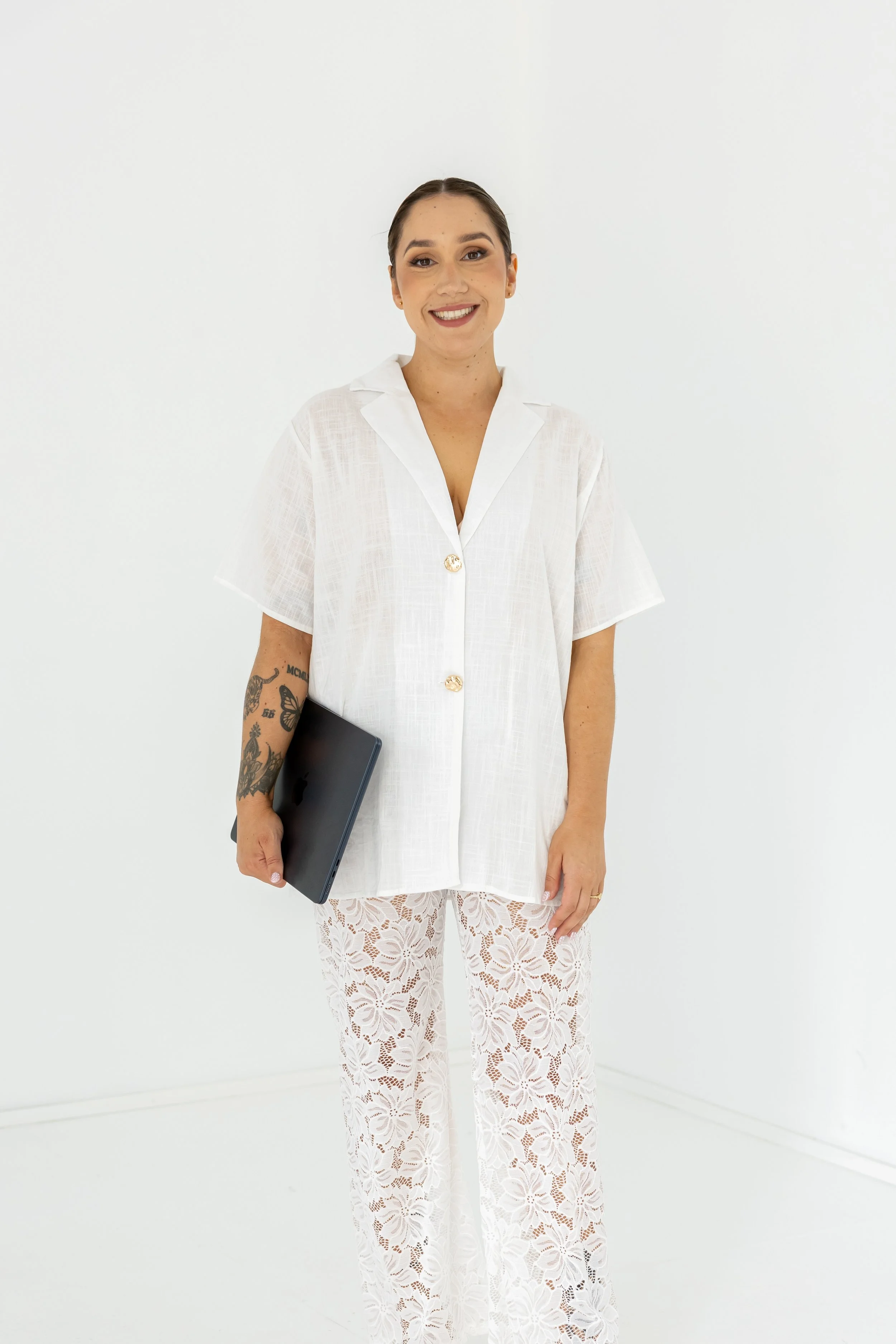 A woman smiling and holding a closed black laptop, wearing a white short-sleeved shirt with gold buttons and white lace pants, standing against a plain white background.