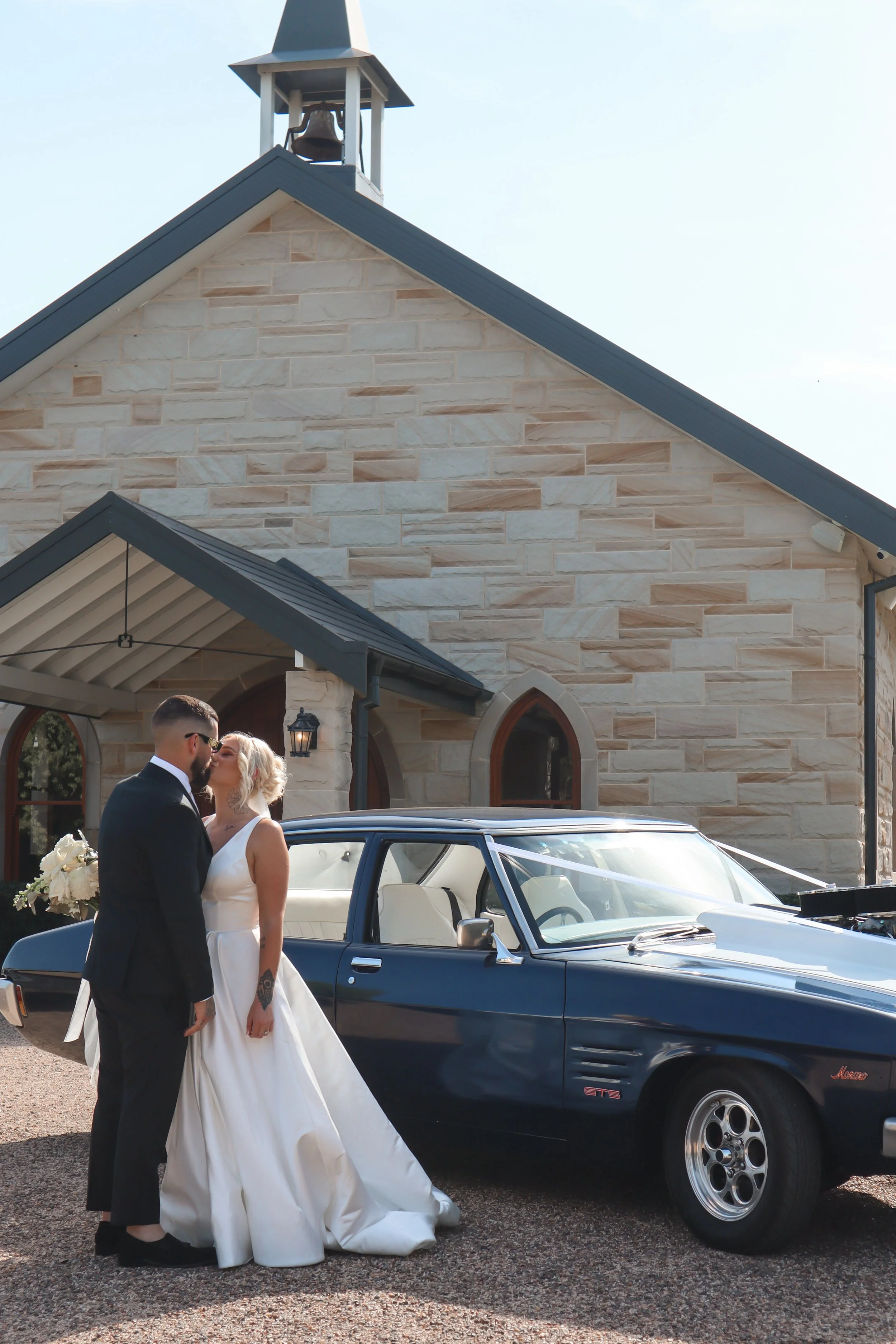 A bride and groom share a kiss in front of a vintage black car outside a stone church with a bell tower