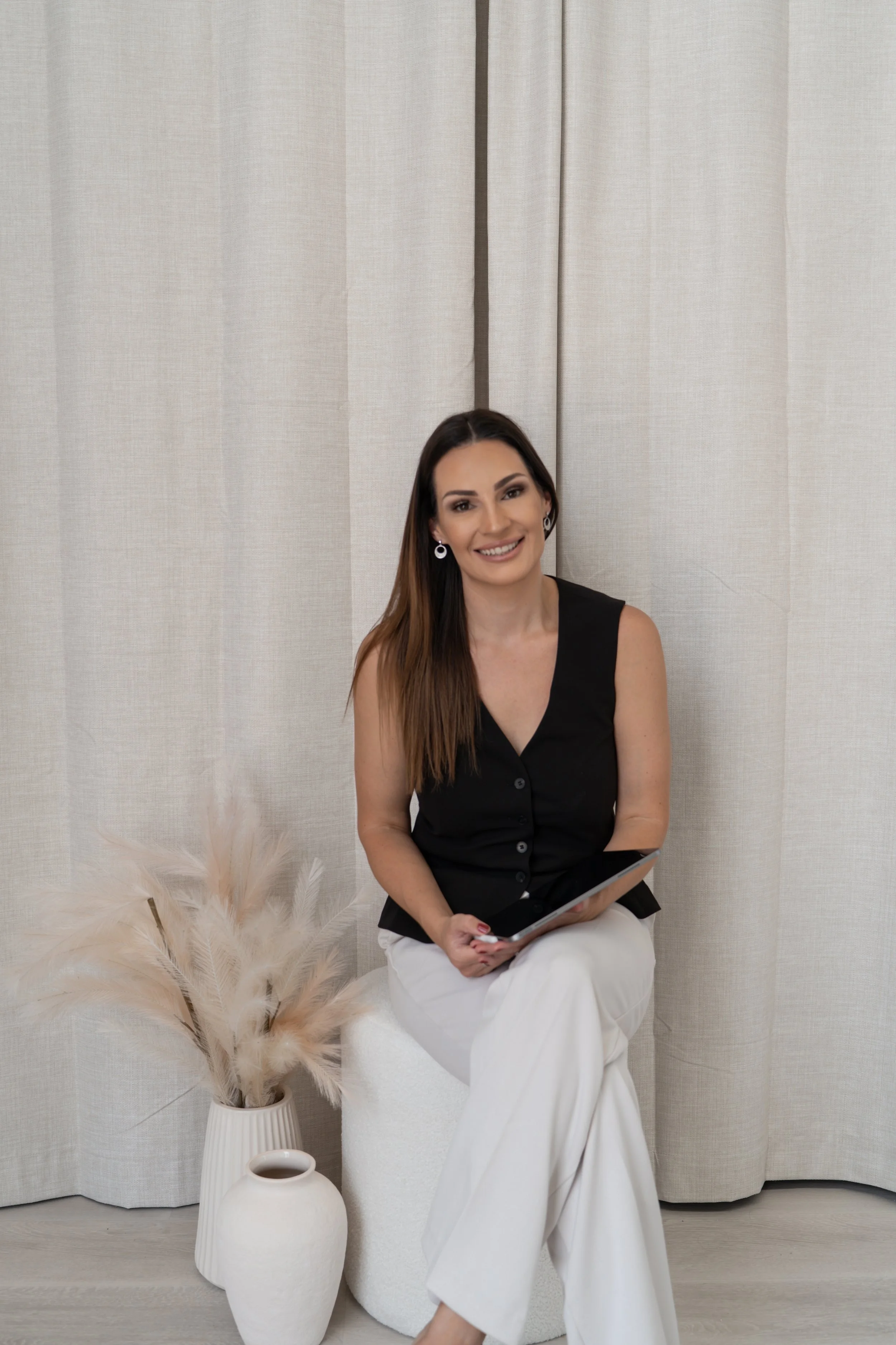A woman with long brown hair, wearing earrings, a black sleeveless top, and white pants, sitting on a white stool, holding a tablet, smiling, with beige curtains and decorative vases with pampas grass nearby.