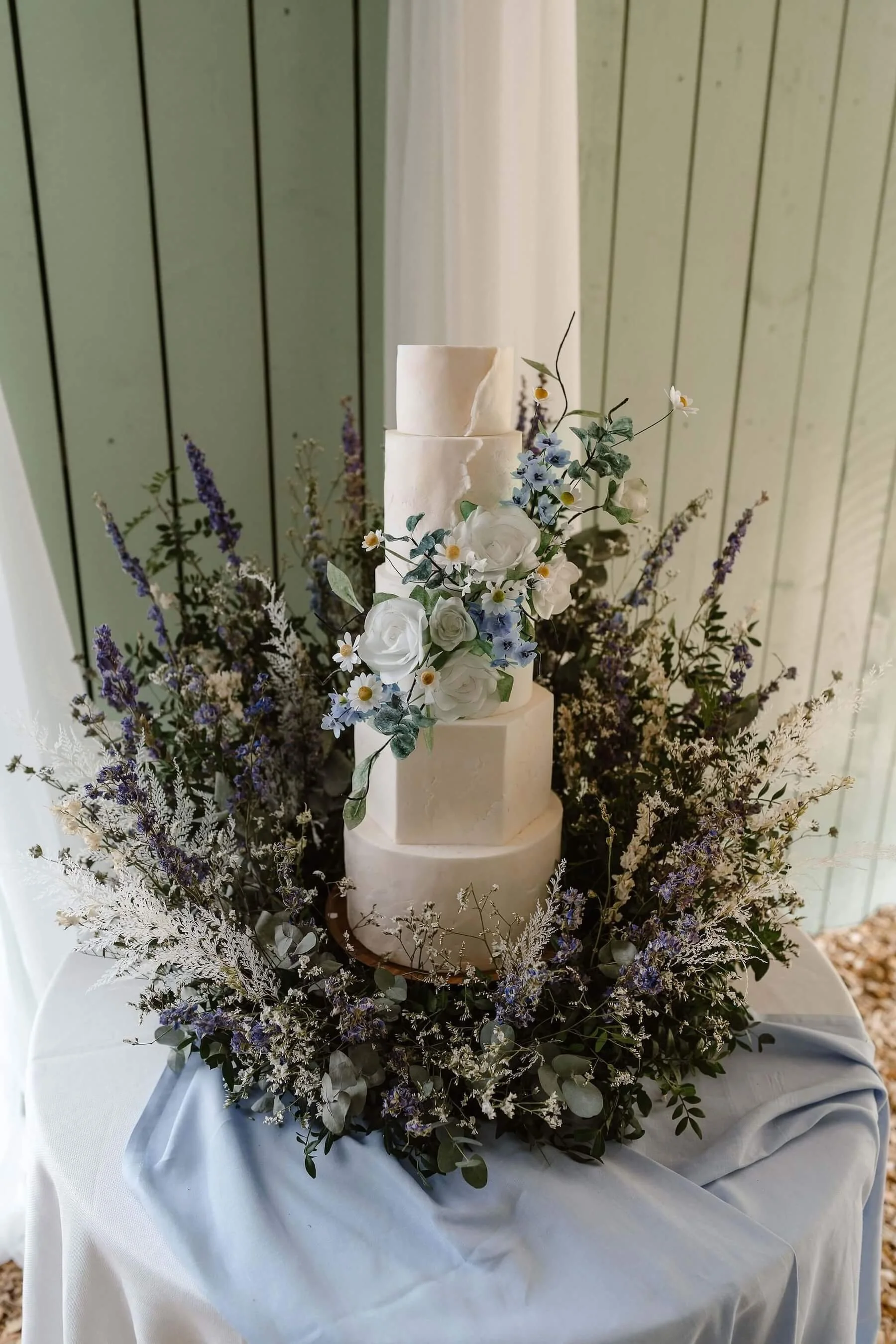 Elegant white wedding cake with floral decorations on a table with a white tablecloth, surrounded by a wreath of purple and white flowers and greenery.