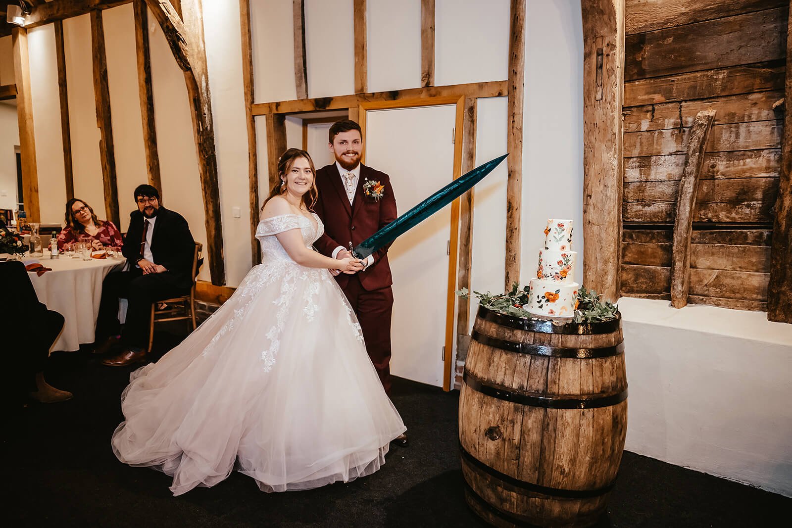 Bride and groom in wedding attire cutting a wedding cake with a large sword, surrounded by wedding guests seated at tables, in a rustic wooden interior.