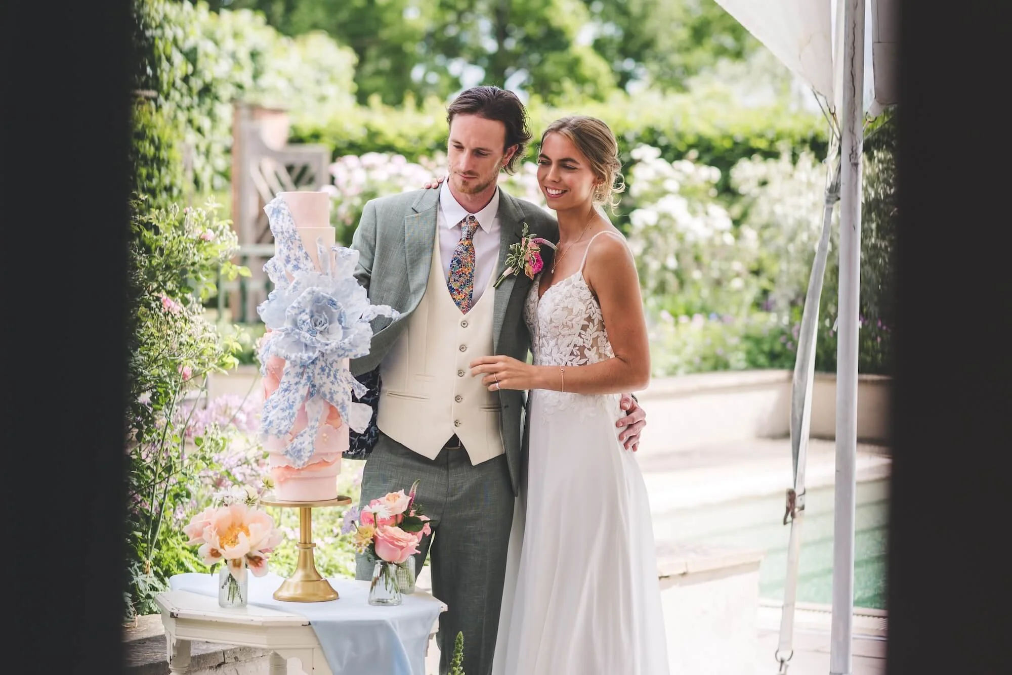 A bride and groom at their reception with a tiered wedding cake on a table, surrounded by flowers and greenery in an outdoor setting.