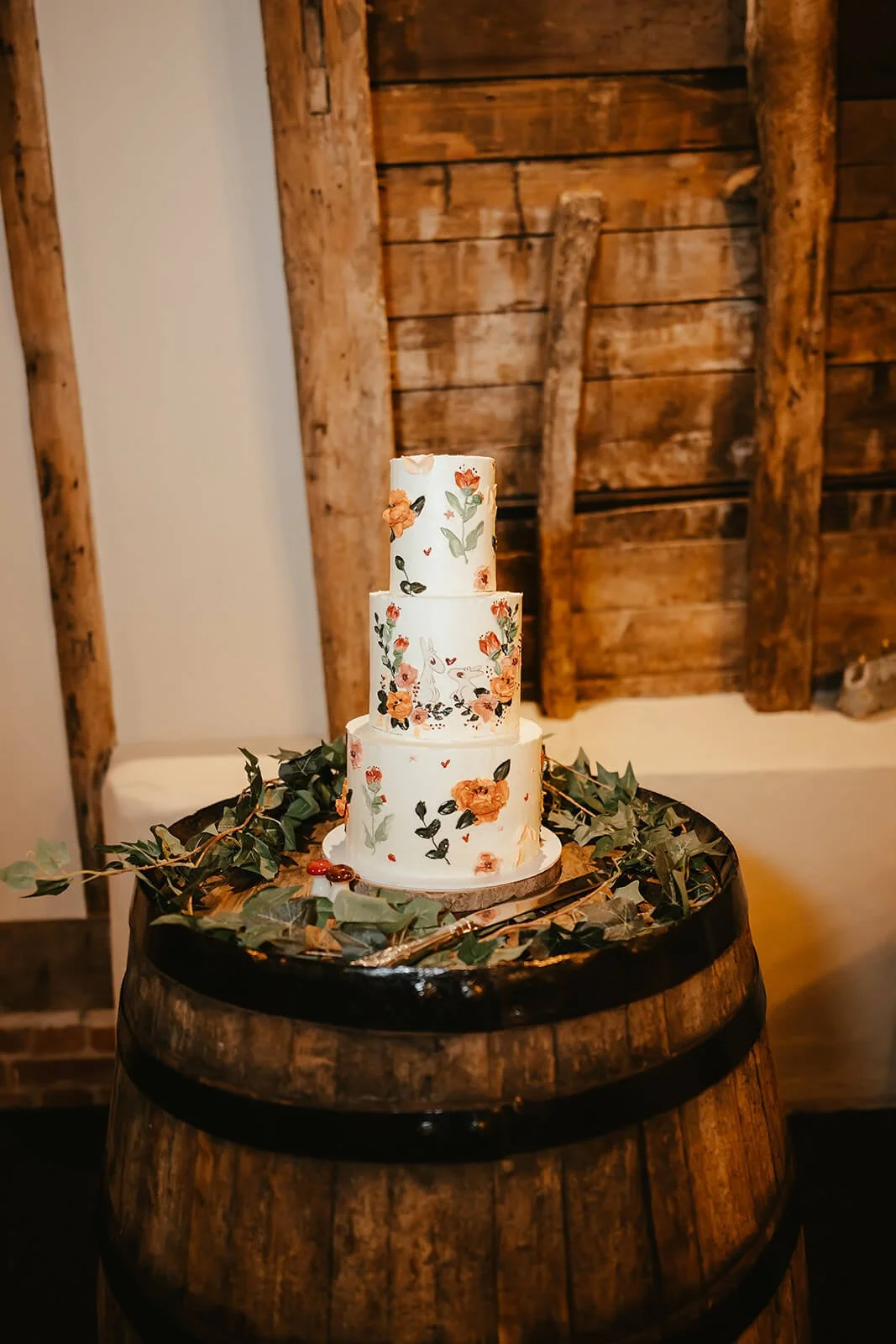 A three-tiered wedding cake decorated with floral and nature-inspired patterns, placed on a wooden barrel surrounded by green foliage, against a rustic wood-paneled wall.