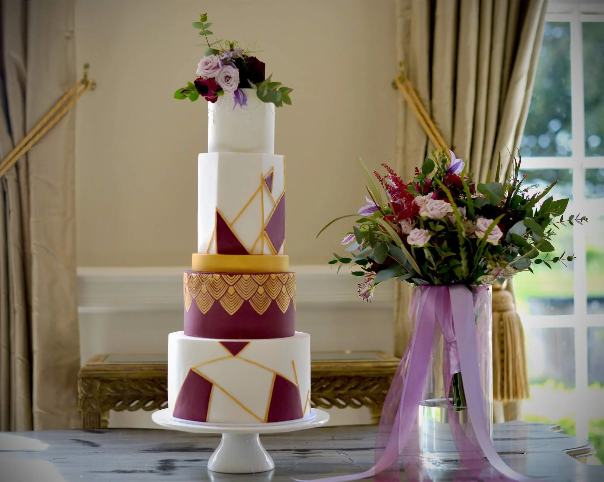 A multi-tiered wedding cake with geometric patterns in burgundy, white, and gold, topped with a bouquet of flowers, placed on a white cake stand