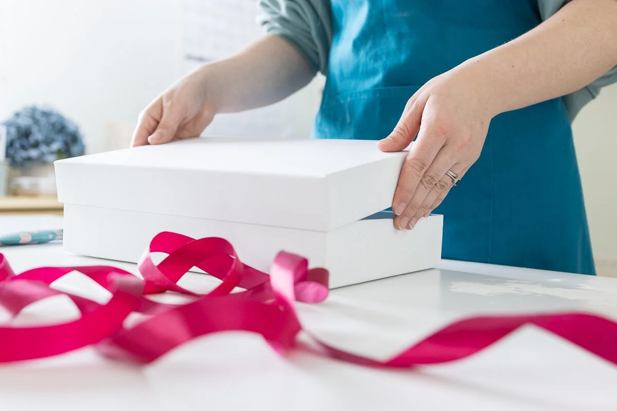 Person opening a white gift box with pink ribbon on a white table.