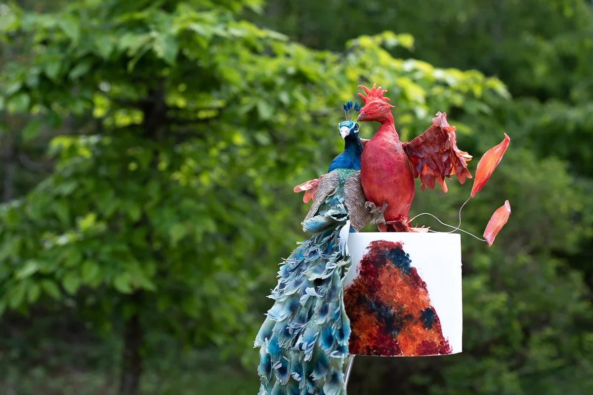 Cake featuring a sculpture of a peacock and a phoenix with abstract and feather details, placed on a white cylindrical base outdoors with green trees in the background.