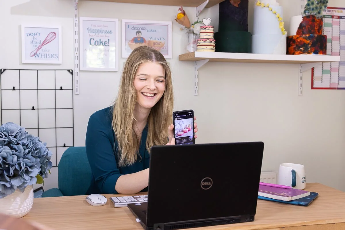 Ruth sitting at a desk in a home office, smiling and holding up her phone to the laptop, as if showing it to someone on a call.