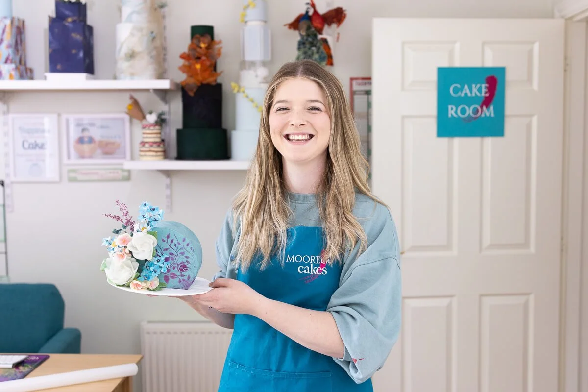 A smiling young woman in a blue apron holding a decorated cake with pink and blue floral decorations inside a bakery or cake shop.