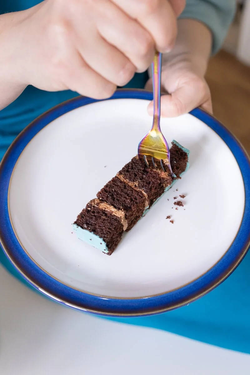 A person holding a fork and taking a piece from a rectangular slice of chocolate cake