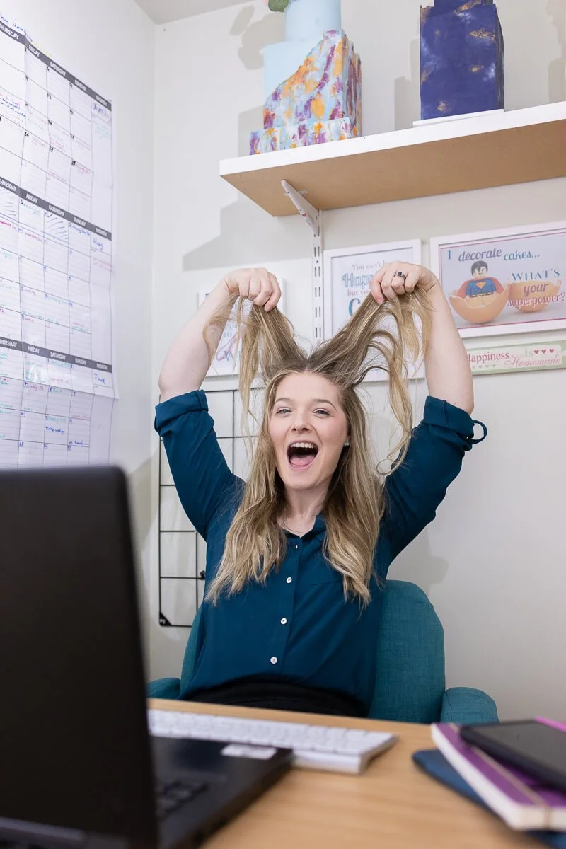 Ruth, a woman with long blonde hair, wearing a blue shirt, sitting at a desk, holding her hair up and smiling at the camera with a laptop and notebooks in front of her, in a bright office
