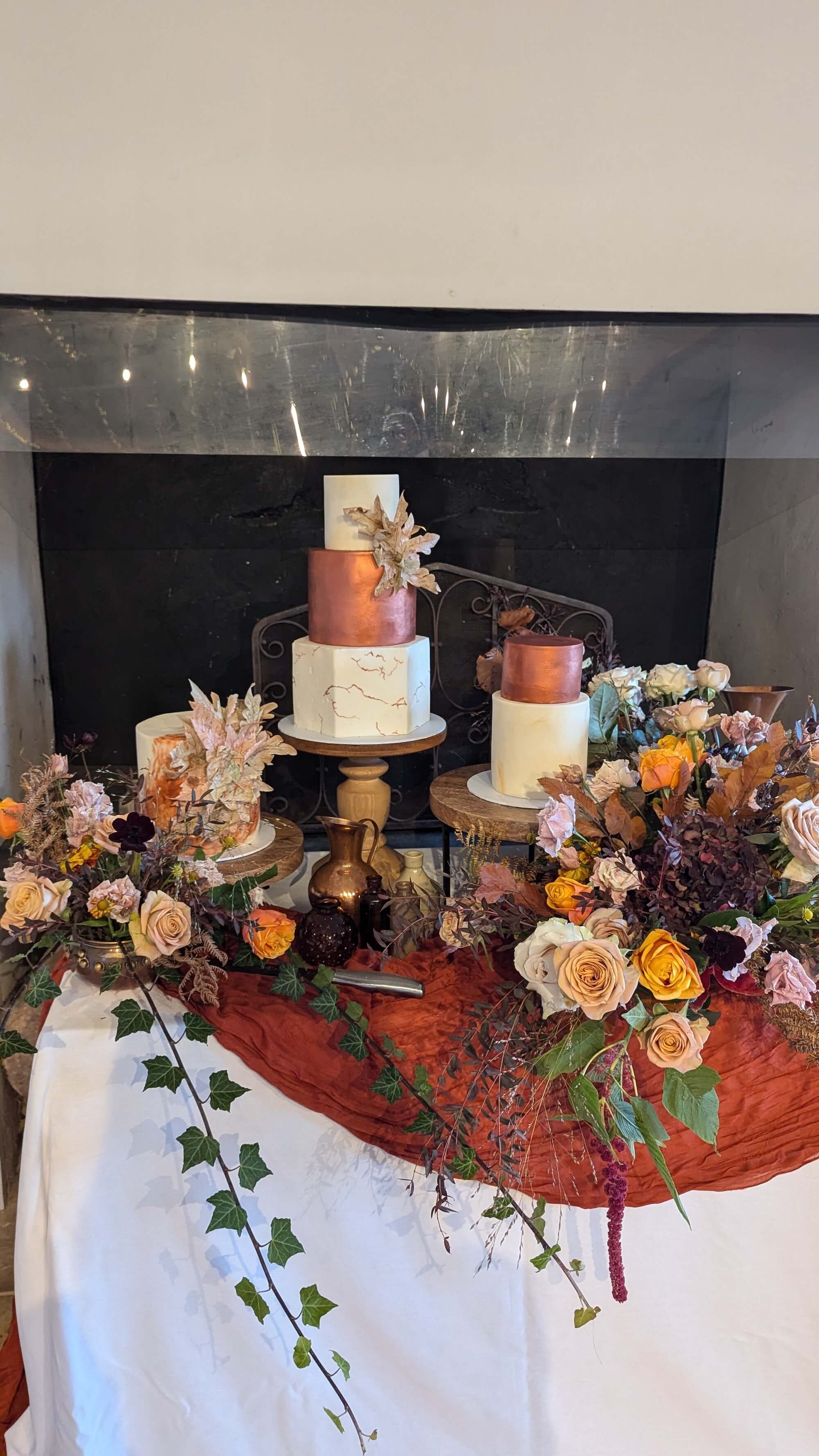 A table decorated with flowers and a burnt orange tablecloth featuring a three-tiered wedding cake with a marbled white and gold design