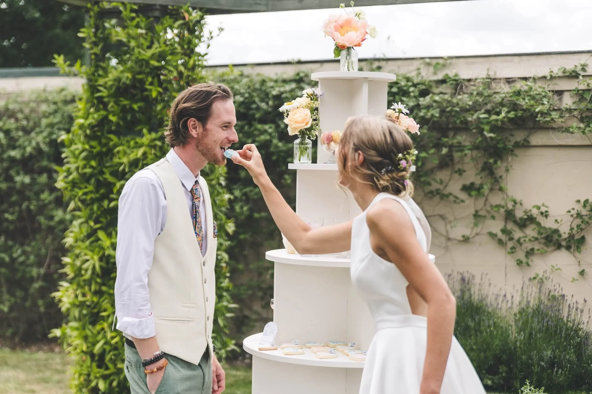 A bride in a white dress feeds a groom a piece of cake next to a dessert stand
