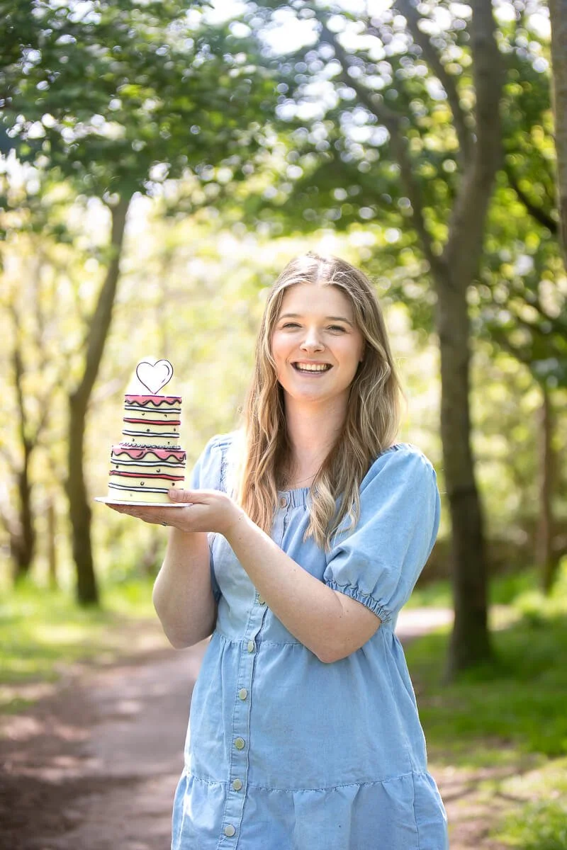 Ruth, a woman with long blonde, hair smiling and holding a small three-tiered cake with a heart decoration on top, outdoors in a wooded park on a sunny day