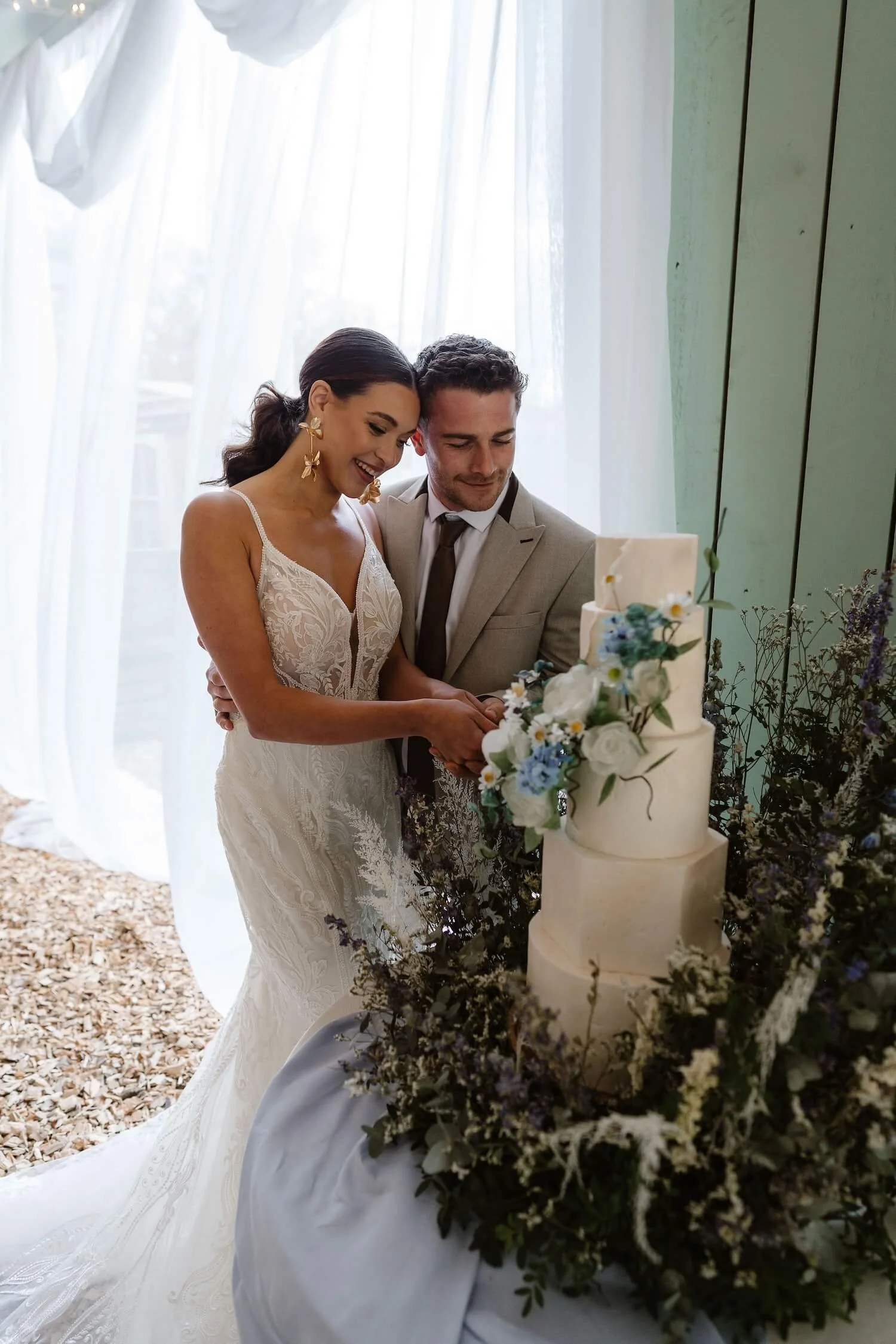 A bride and groom cutting a wedding cake together, surrounded by floral decorations, in a bright room with sheer curtains.