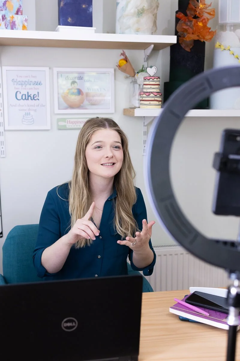 Ruth Moore talks in front of a phone in a ring light, in a decorated office space with framed posters, artwork, and decorative items on shelves