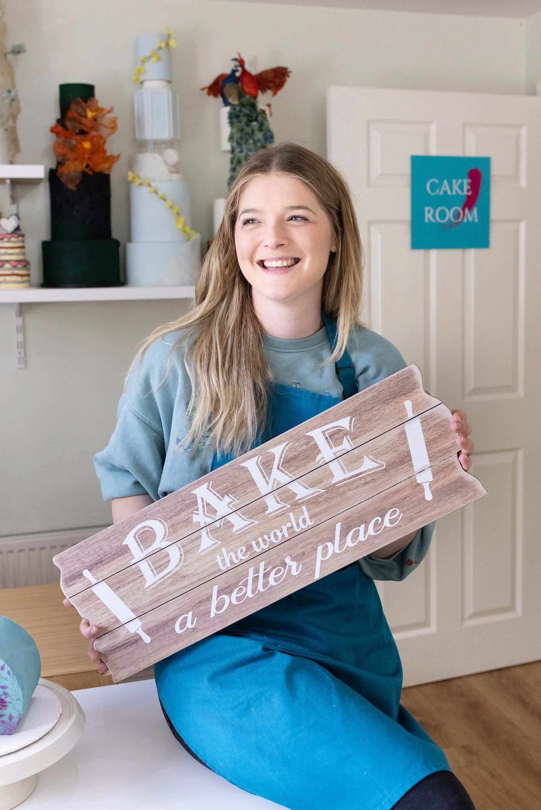 Ruth perched on a table holding a wooden sign that reads 'Bake the world a better place'