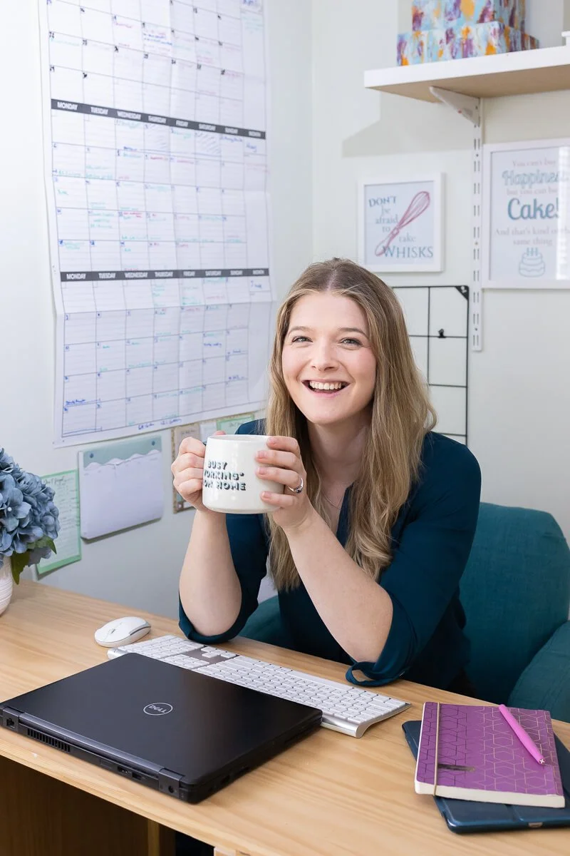 Ruth sitting at a desk holding a white coffee mug, smiling, with a large calendar on the wall behind her