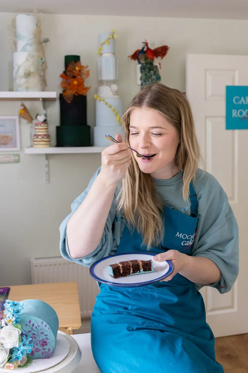 Ruth tastes a slice of cake. She is holding a plate with a piece of chocolate cake and using a fork to eat it.