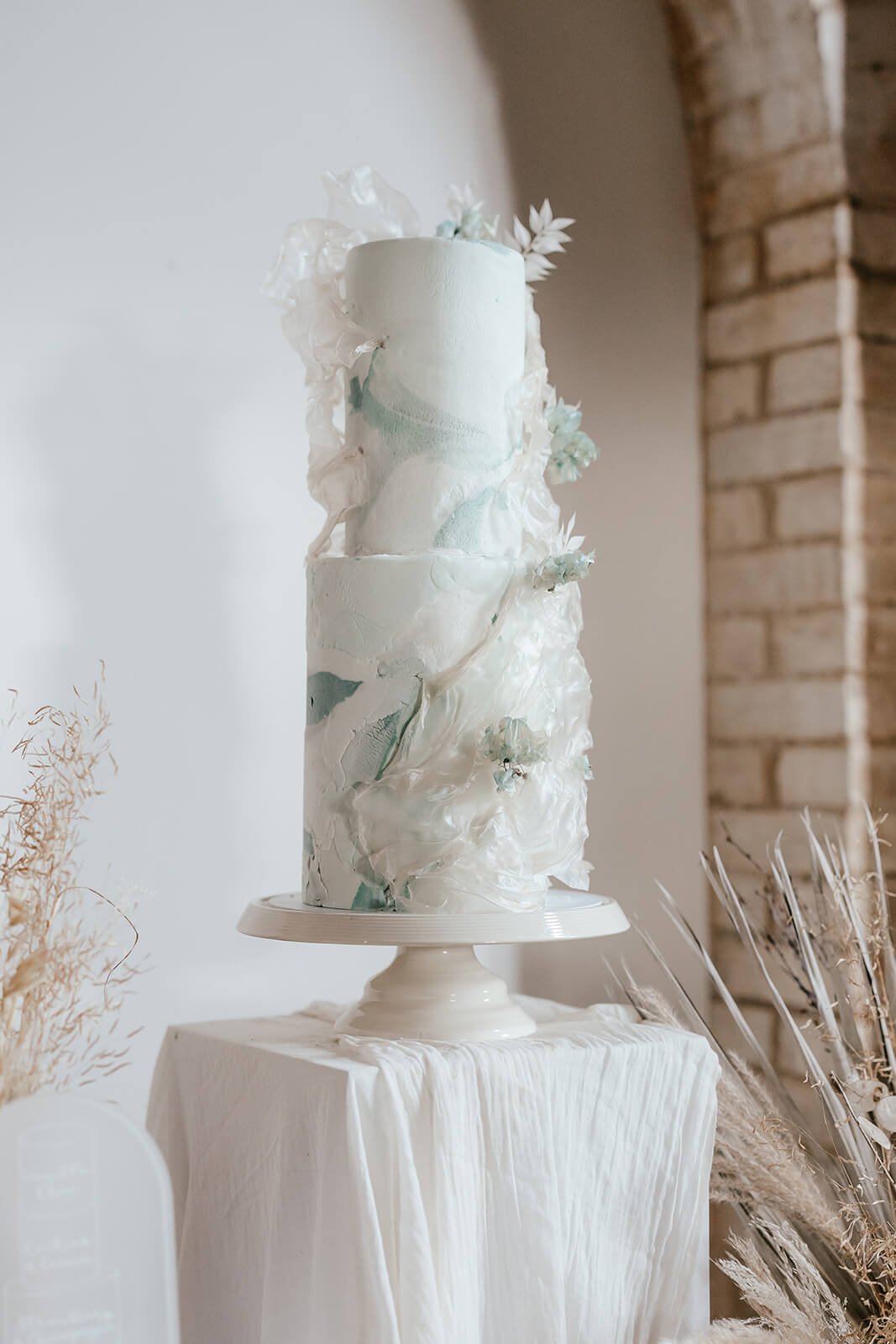 A tall, white, three-tier wedding cake decorated with white and light blue abstract floral and leaf details