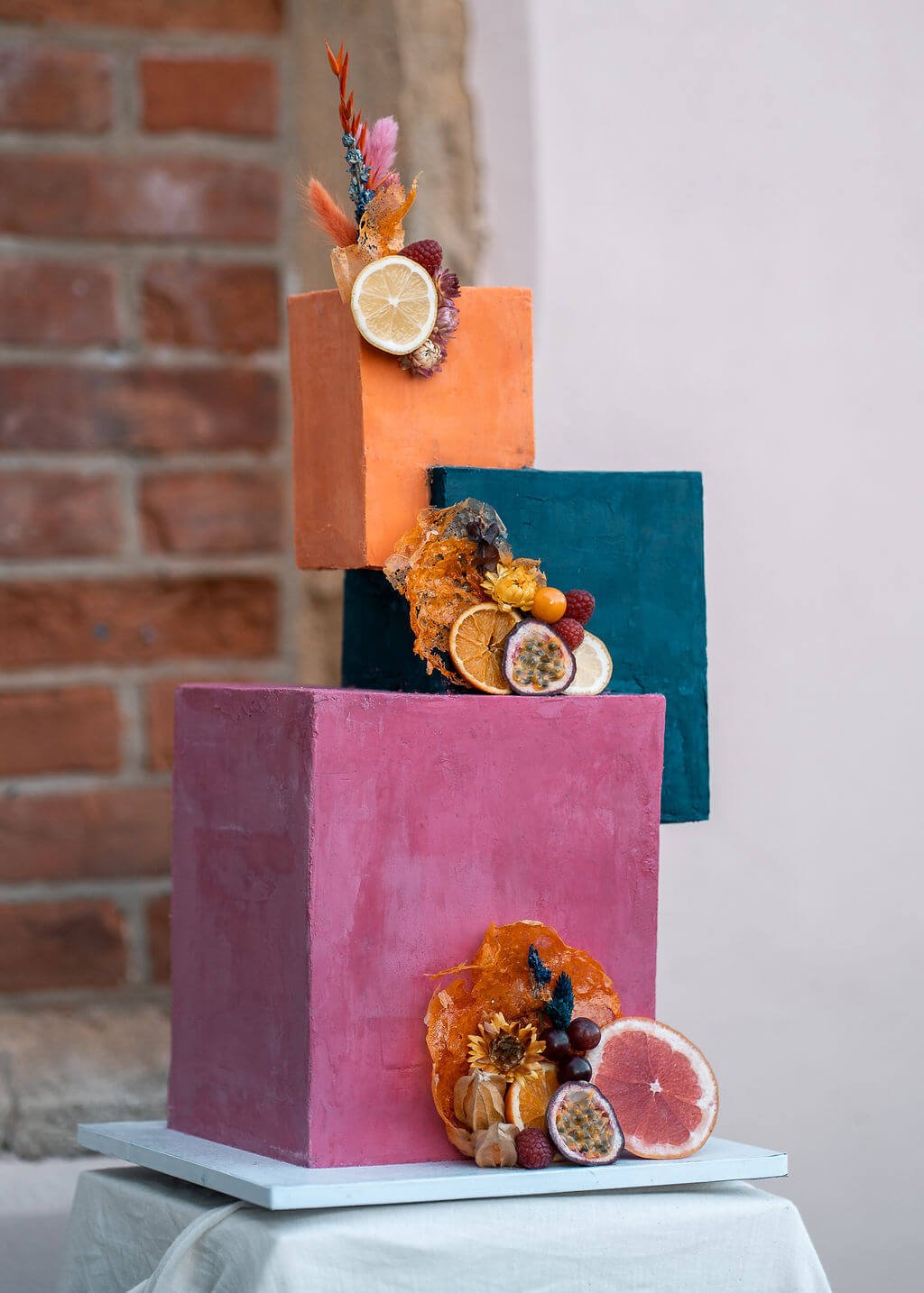 A three-tiered wedding cake with each tier painted in different colors: pink, blue, and orange. The cake is decorated with slices of citrus fruits, berries, dried flowers, and other edible adornments