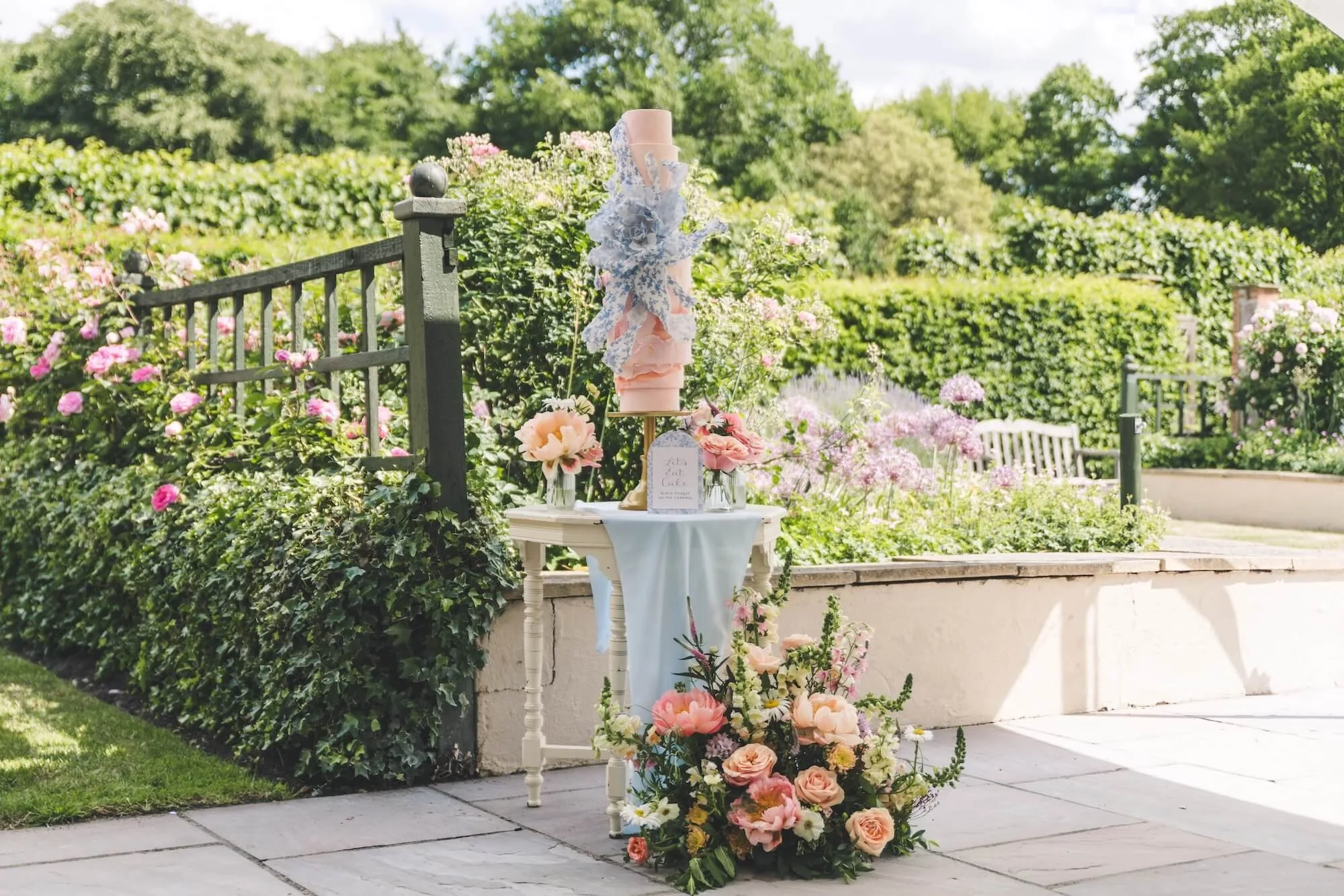 An outdoor floral display with floral and lace tiered wedding cake, in a garden setting