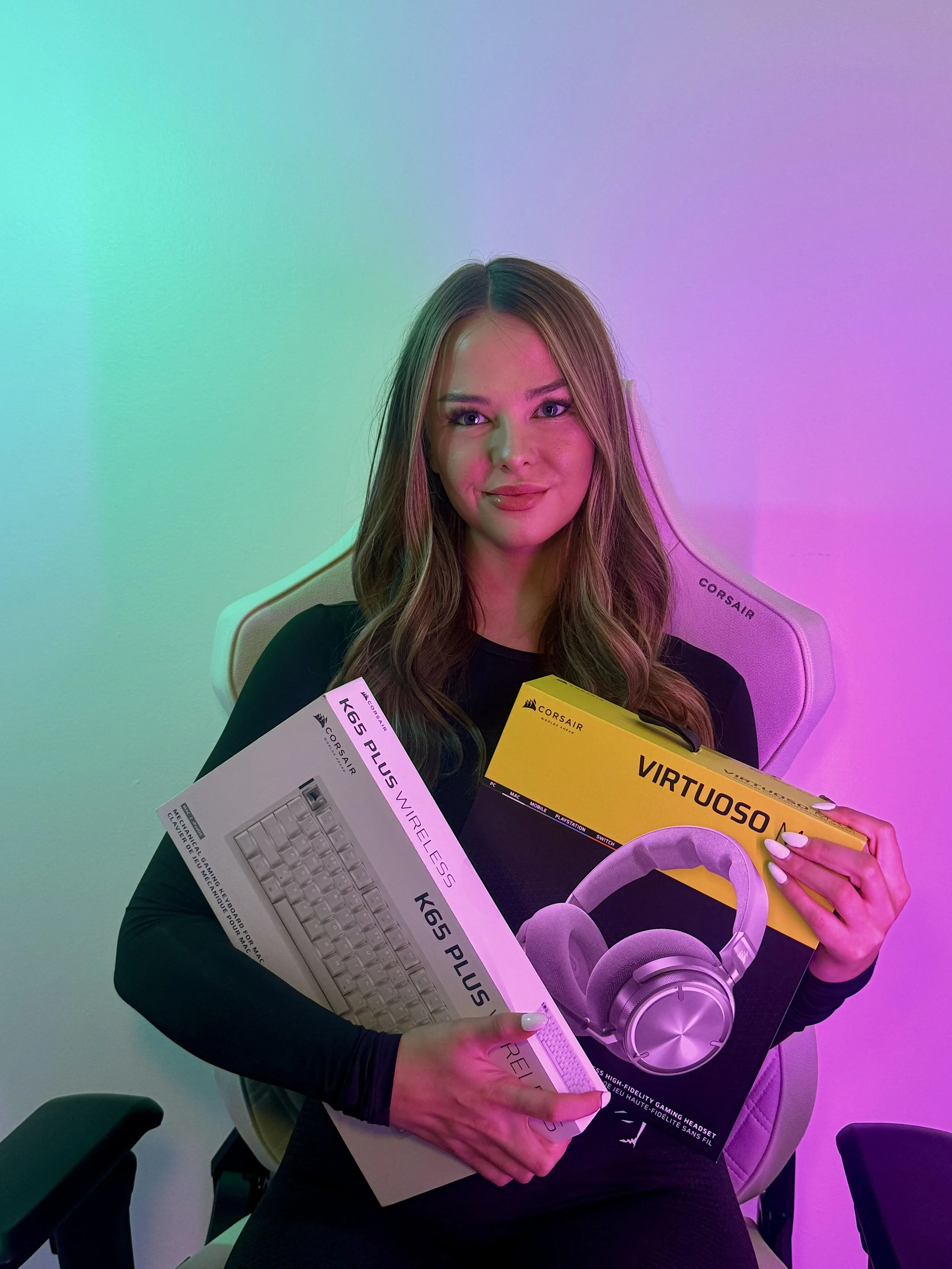A woman sitting in a white Corsair gaming chair holding two boxed computer accessories: a mechanical keyboard and a headset, with colored lighting in the background.