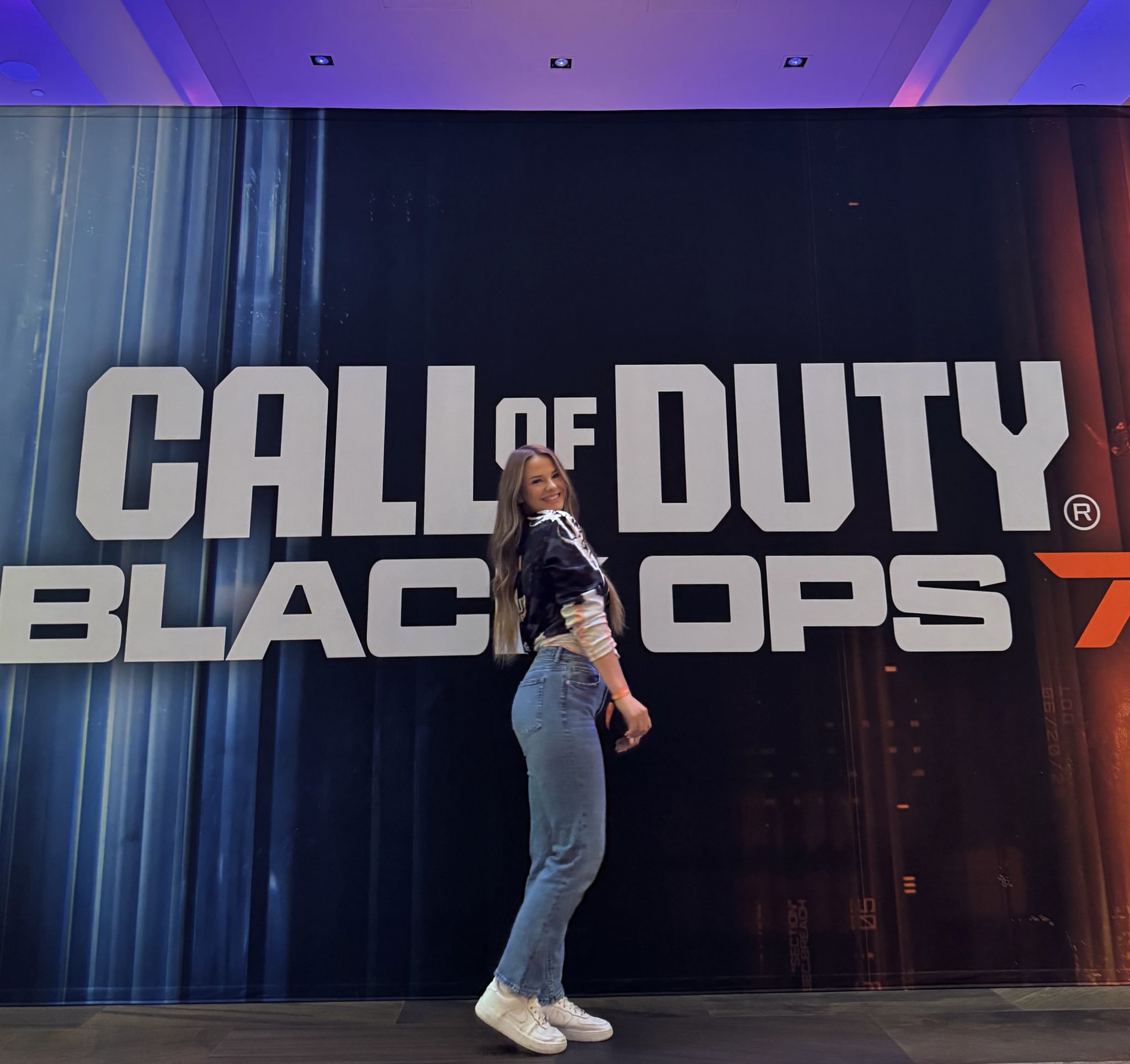 A woman standing in front of a large Call of Duty: Black Ops 4 poster, smiling and posing sideways.