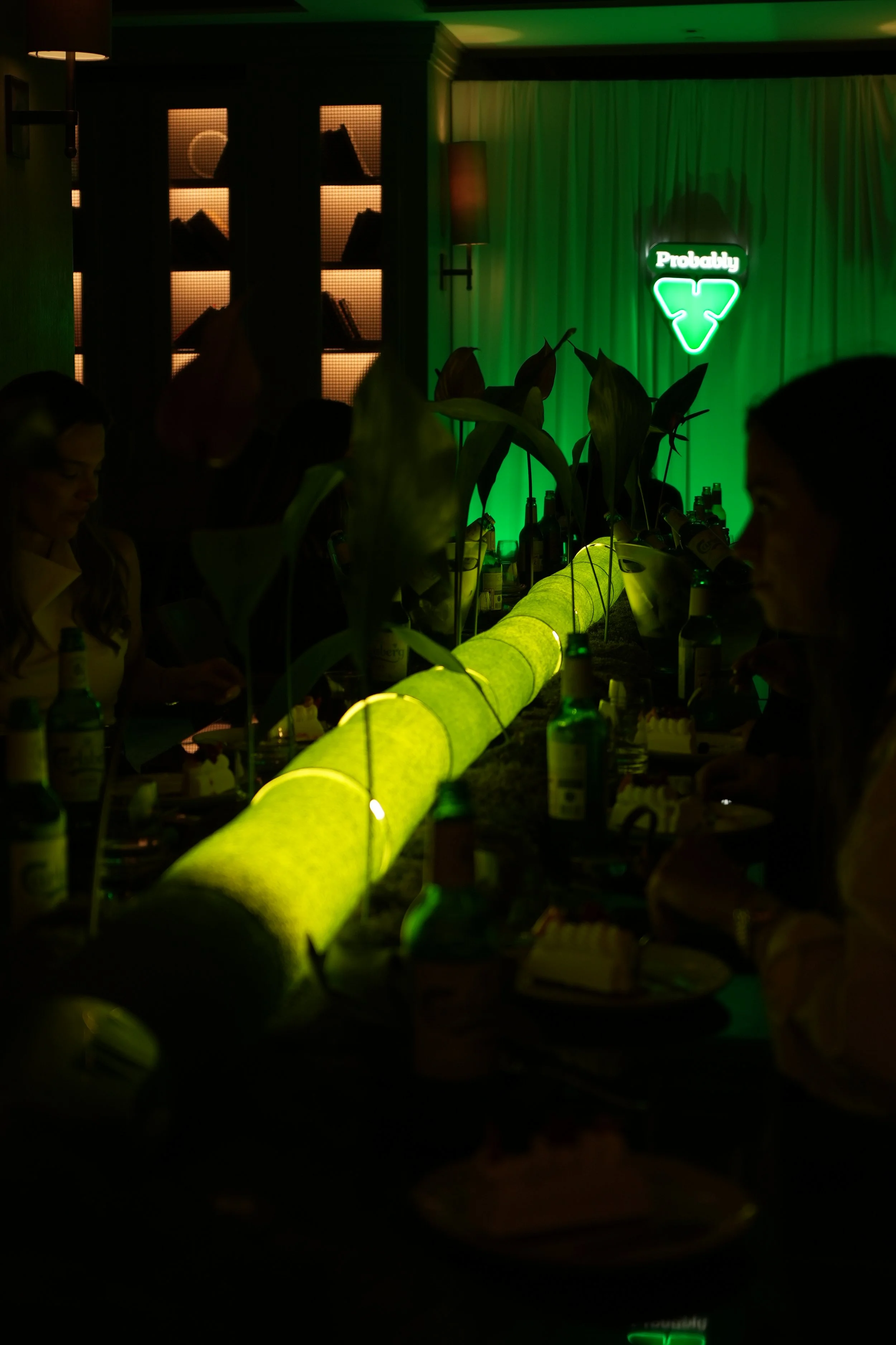 Dimly lit bar with a glowing green bar top, plants, and a neon sign that says 'Probably' with a recycling symbol, with patrons sitting around the bar.