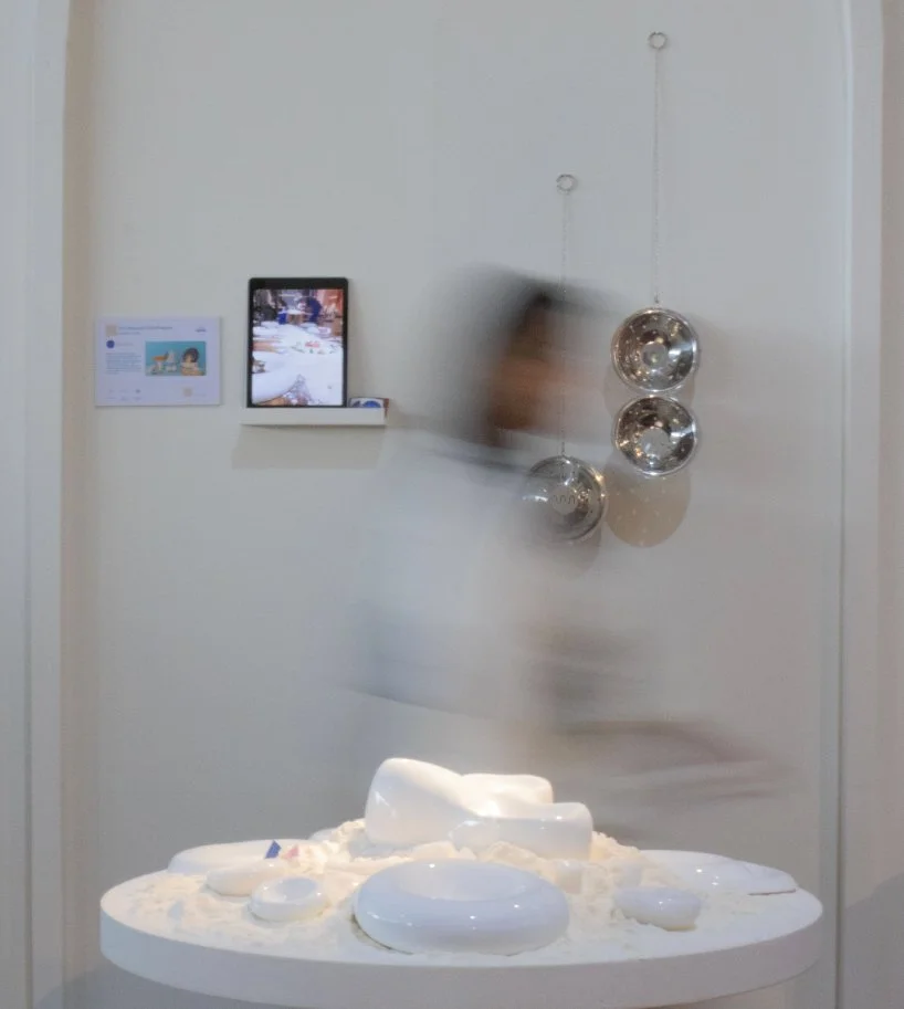 A display with a white surface covered in white material, with various white ceramic objects on top, including bowls and a pitcher. In the background, a wall-mounted screen, some hanging metallic spherical objects, and a blurred figure are visible.