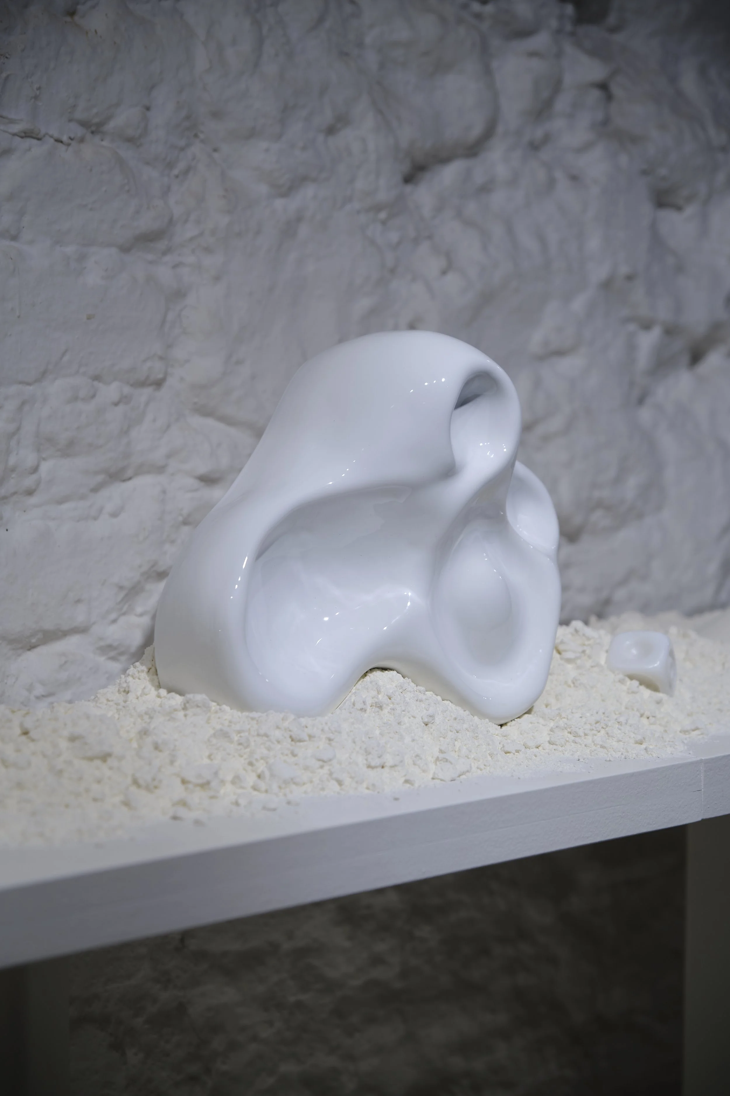 A glossy white abstract sculpture resembling a smooth, curvy, and hollow form, placed on a bed of white sand against a textured white brick wall.