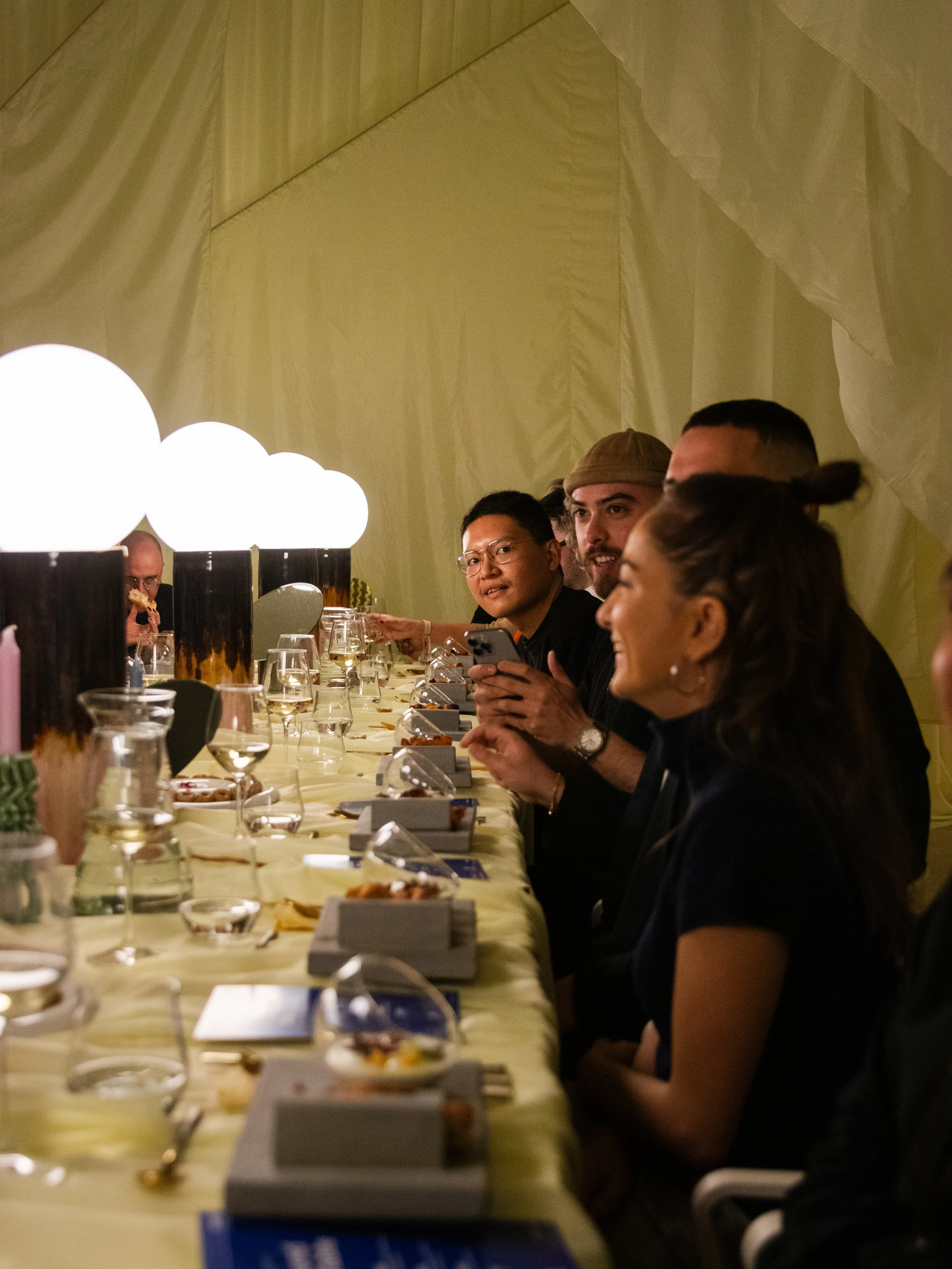 People sitting at a long table during a dinner event, chatting and enjoying food and drinks in a decorated room.