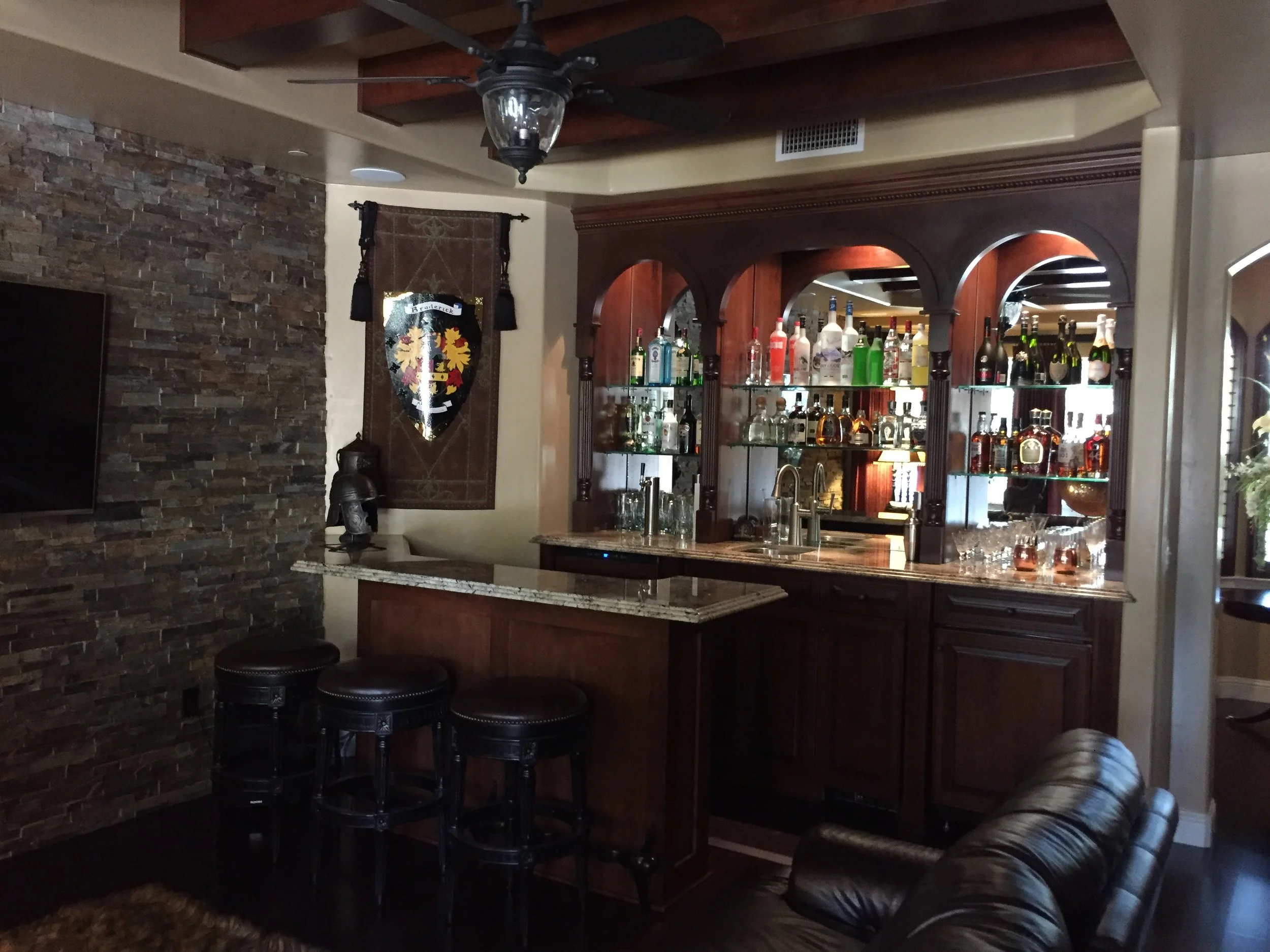 A home bar area with a wooden cabinet filled with various bottles of alcohol, a granite countertop, and three black leather bar stools. The space features a leather couch, a stone accent wall, and a medieval shield with a coat of arms hanging on the 