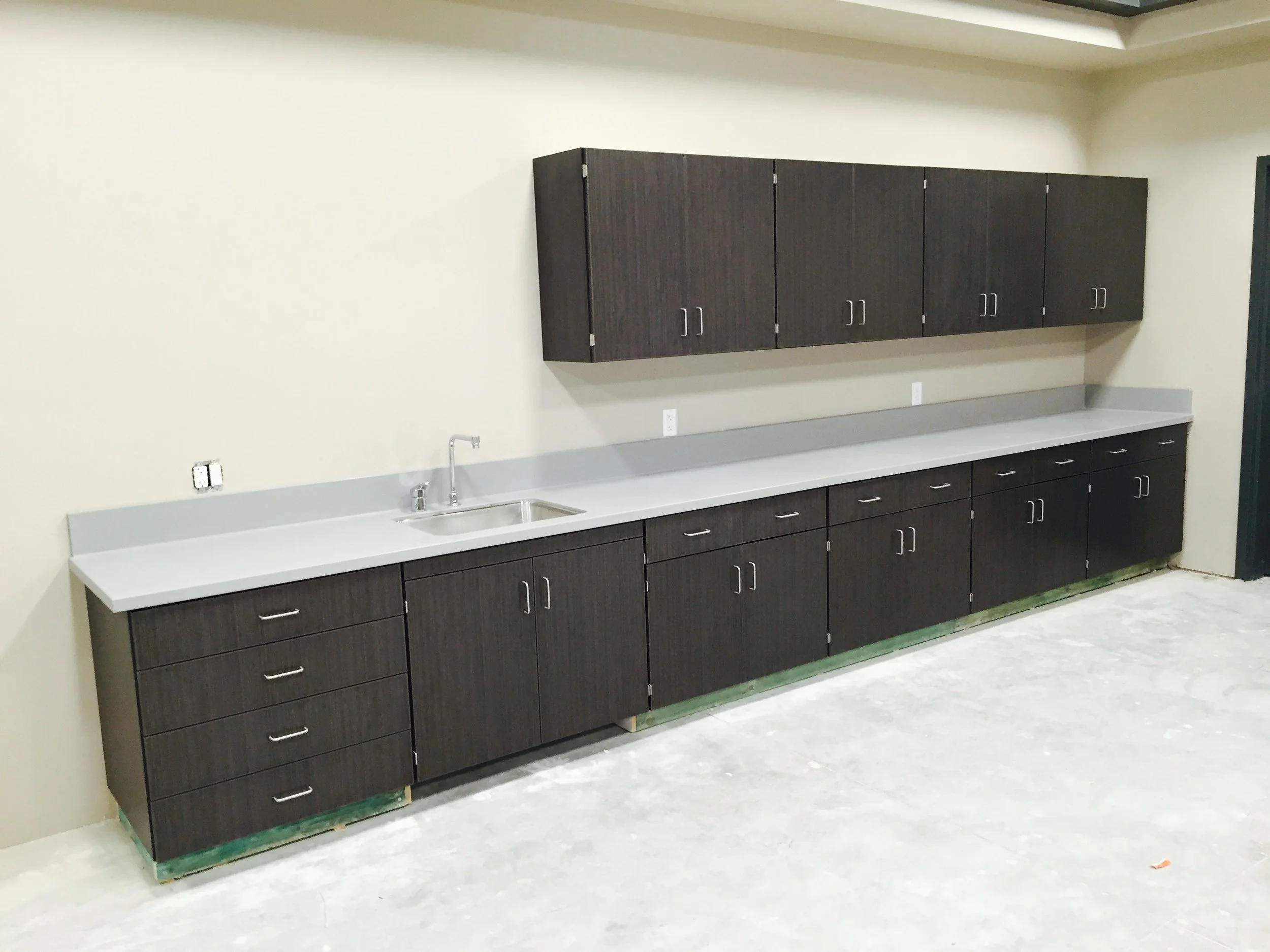 Long countertop with dark wood cabinets above and below, a stainless steel sink with a faucet, and beige walls.