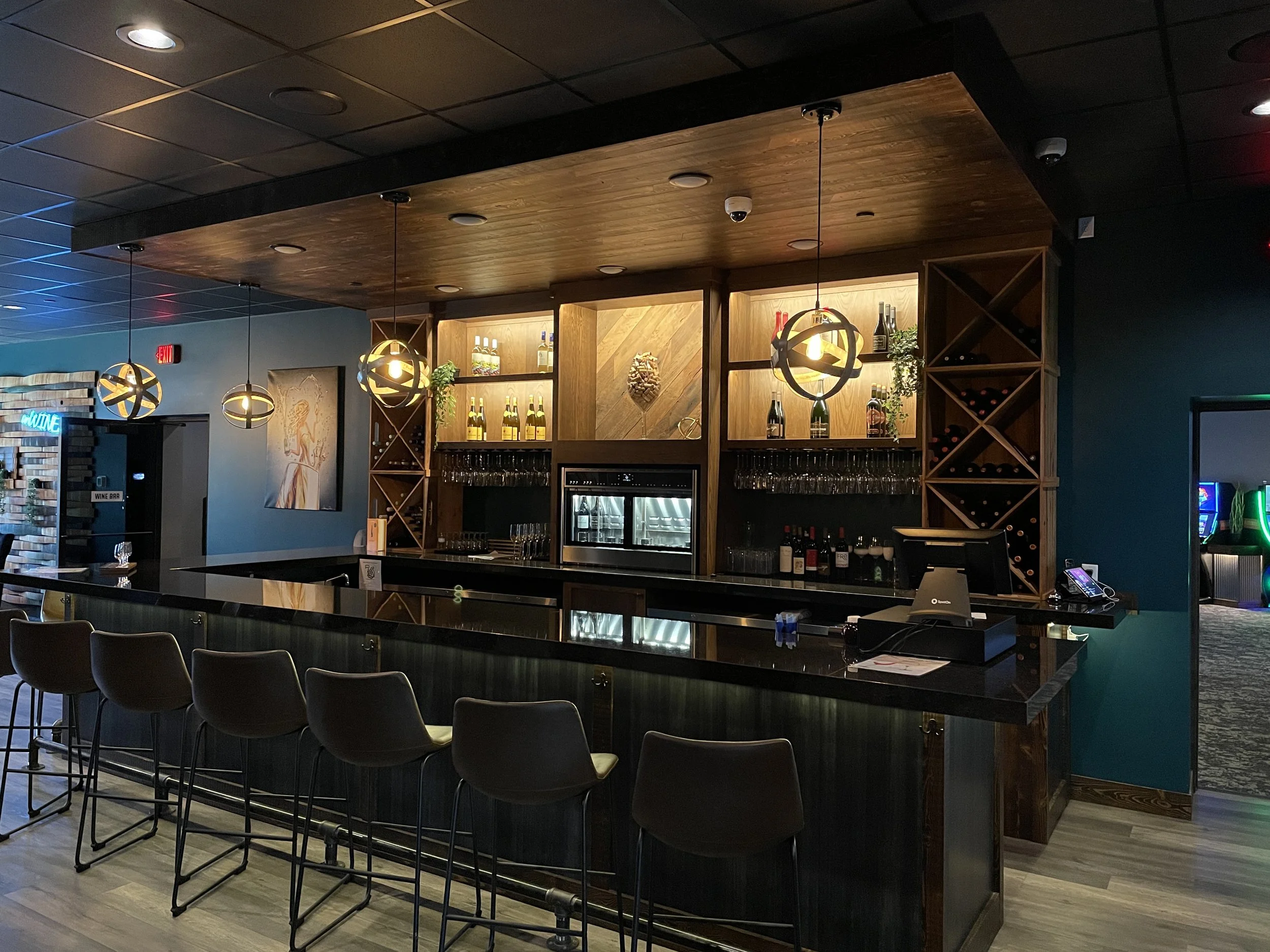 A modern bar with a black countertop, several bar stools, and illuminated wooden shelves holding bottles of alcohol. Three pendant lights hang above the bar, and there is a neon sign to the left. A painting is on the blue wall, and a doorway with a '
