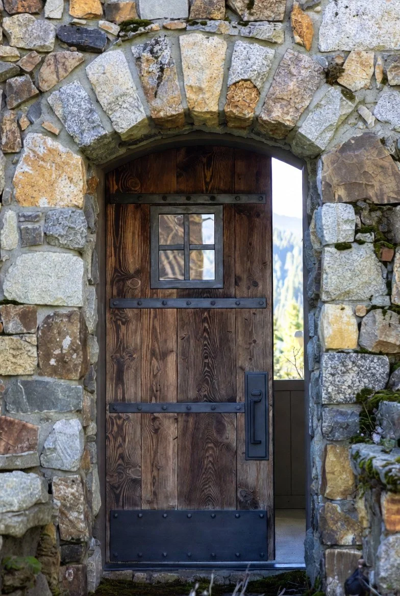 A rustic stone archway leading to a wooden door with black metal hardware and a small window, set in a stone wall.