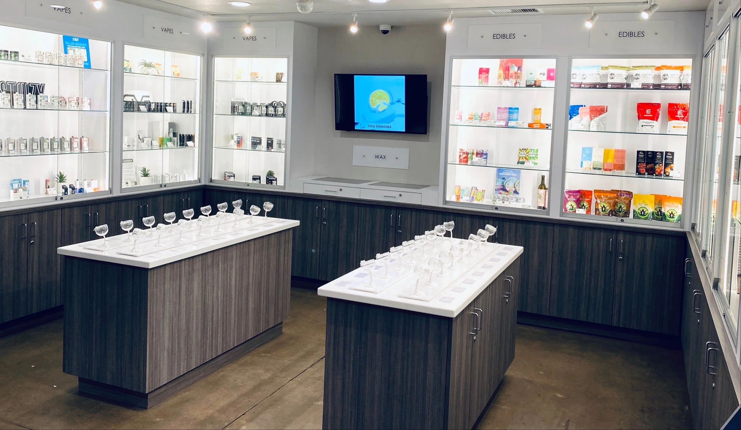 Display of cannabis and edible products in a retail store with glass shelves, two tables with glass samples, and a digital screen on the wall.