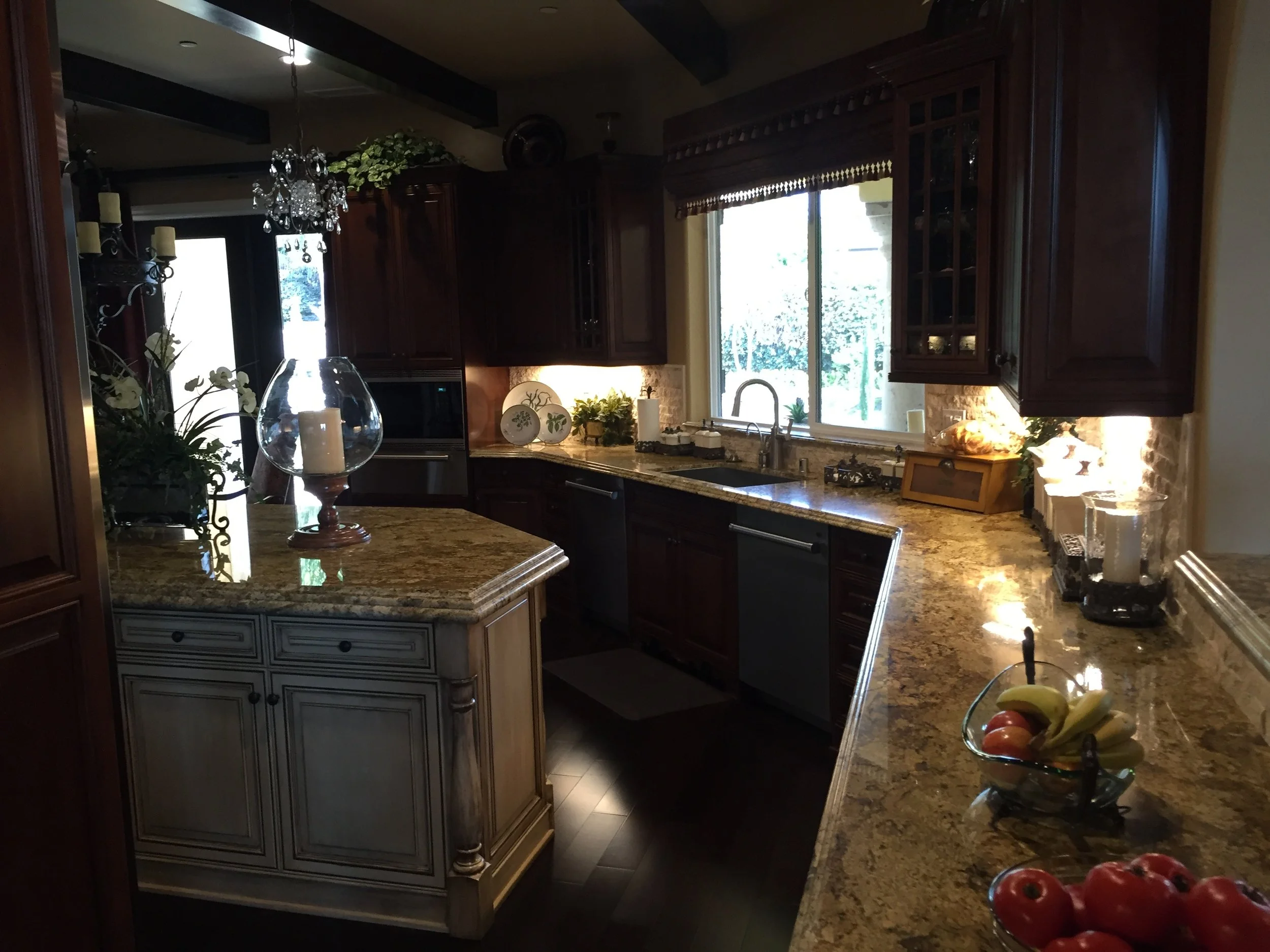 A kitchen with dark wood cabinets, granite countertops, and a window above the sink letting in natural light. Decor includes candles, plants, and fruit bowls.
