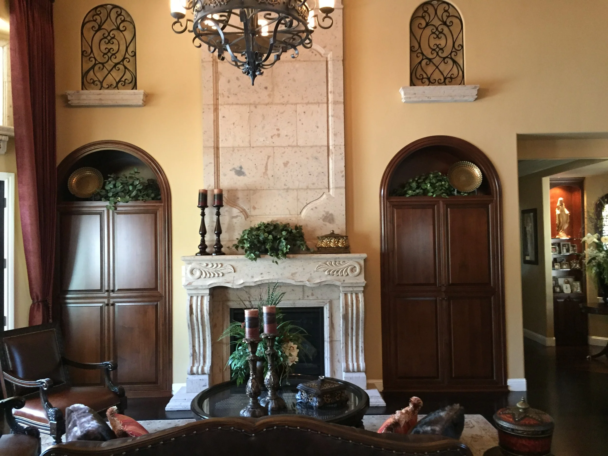 A living room featuring a beige stone fireplace with intricate carved details, topped with green plants and decorative candles. Flanking the fireplace are two dark wooden cabinets with arched tops, each holding decorative plates and greenery. A chand