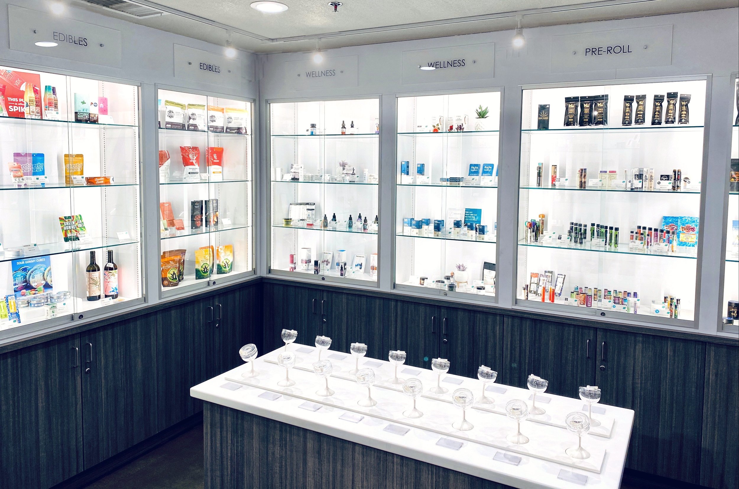 Display case with various health and wellness products, labeled as 'Edibles,' 'Wellness,' and 'Pre-Roll,' in a bright, modern store with glass shelves and a white countertop with empty glass bowls.