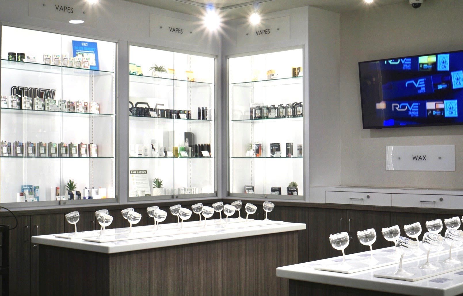 Vape shop display with glass shelves holding various e-cigarette products, a wall-mounted screen, and a white counter with glass pieces for tasting or demonstration.