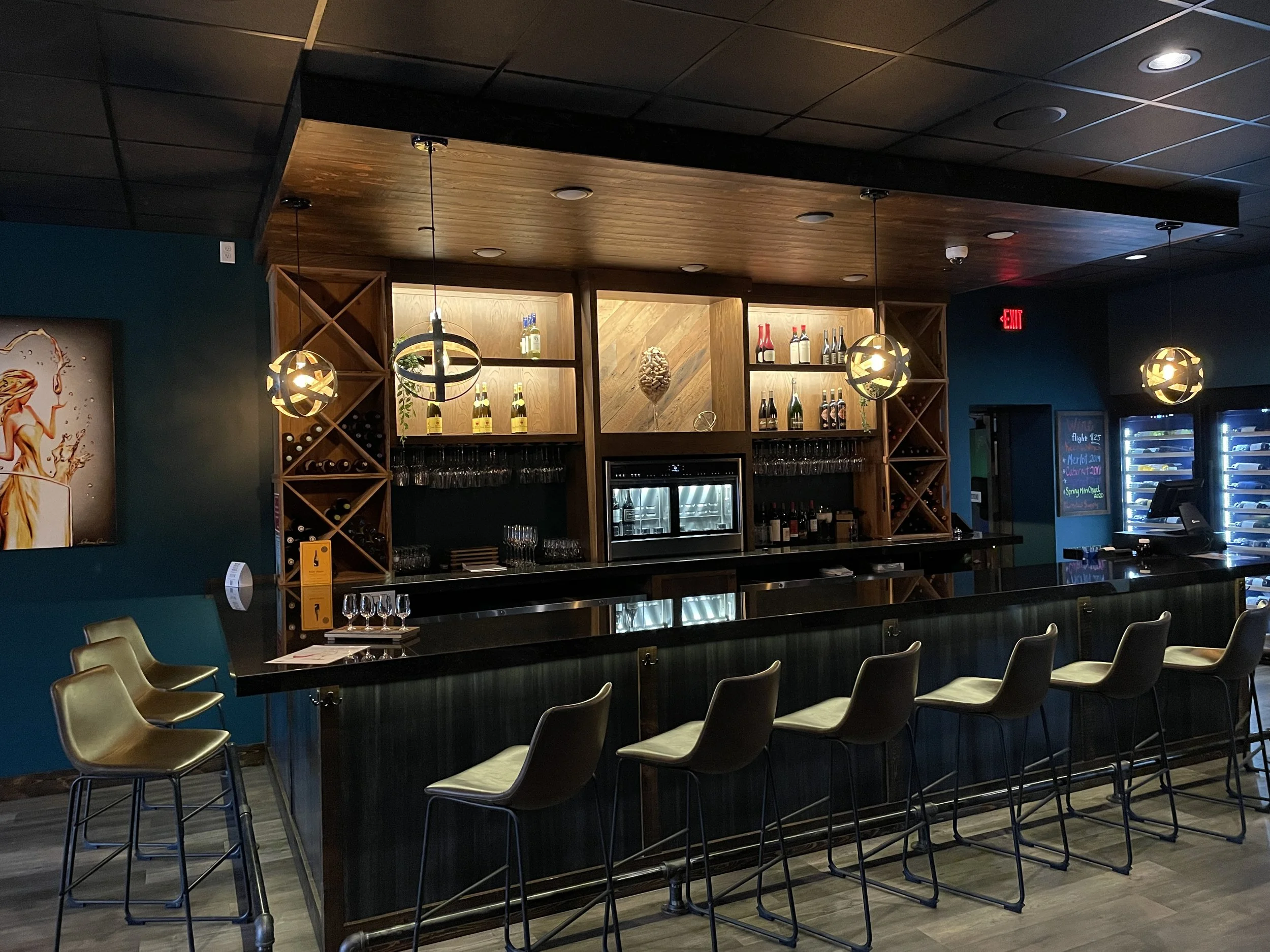 Empty bar with six beige chairs lined up at the black countertop, liquor bottles and glasses on shelves behind the bar, three hanging pendant lights, and a vending machine with drinks to the right.